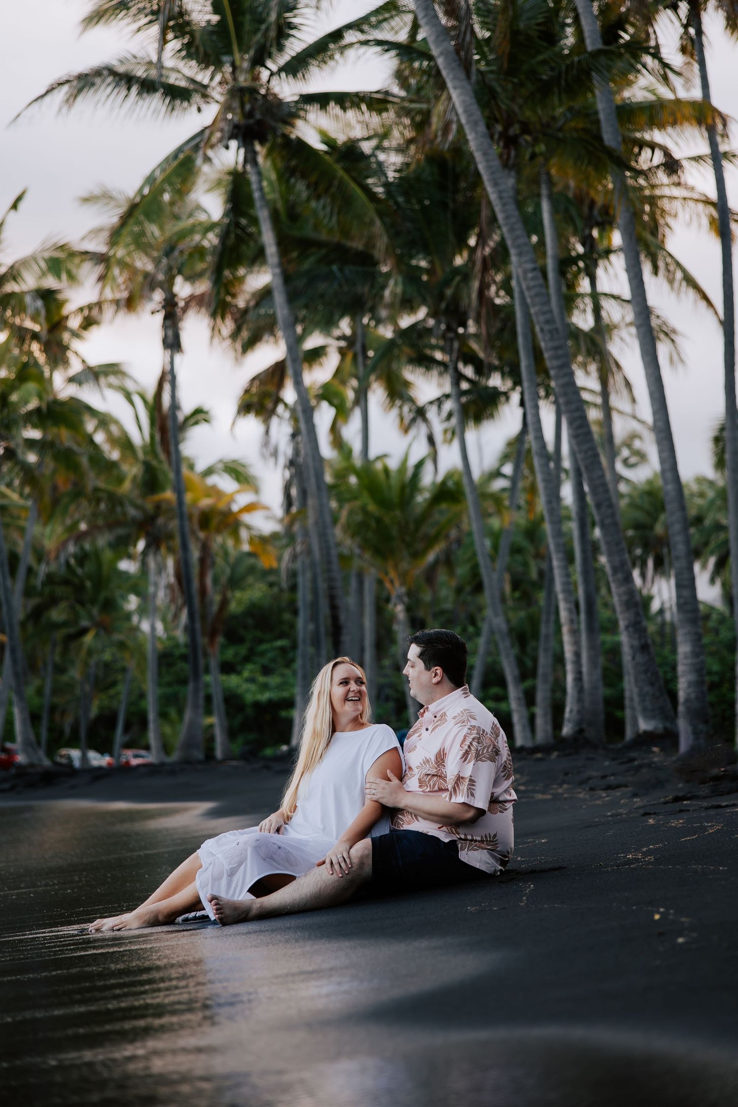 Couple sitting together on black sand beach surrounded by tall palm trees at sunset in Hawaii.