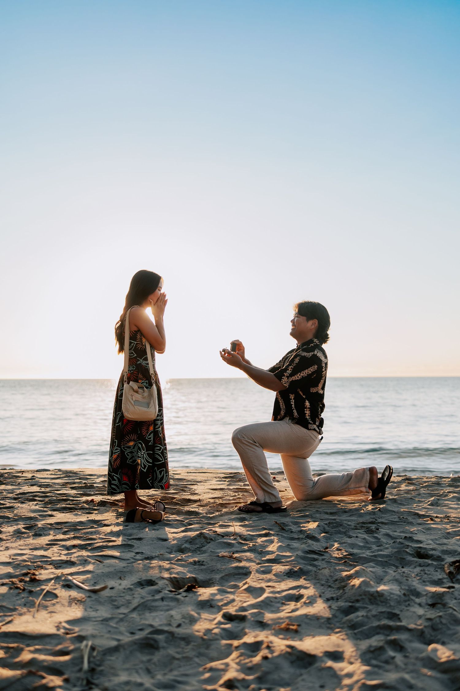 Man proposing to woman on a sandy beach at sunset, kneeling with a ring box as she covers her mouth in surprise.