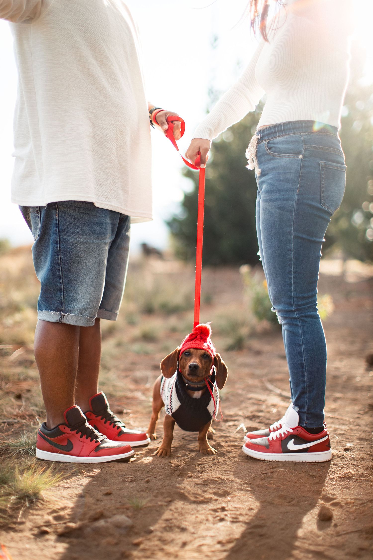 Couple walking a dachshund on a red leash, both wearing matching red and white Nike sneakers on a dirt path.