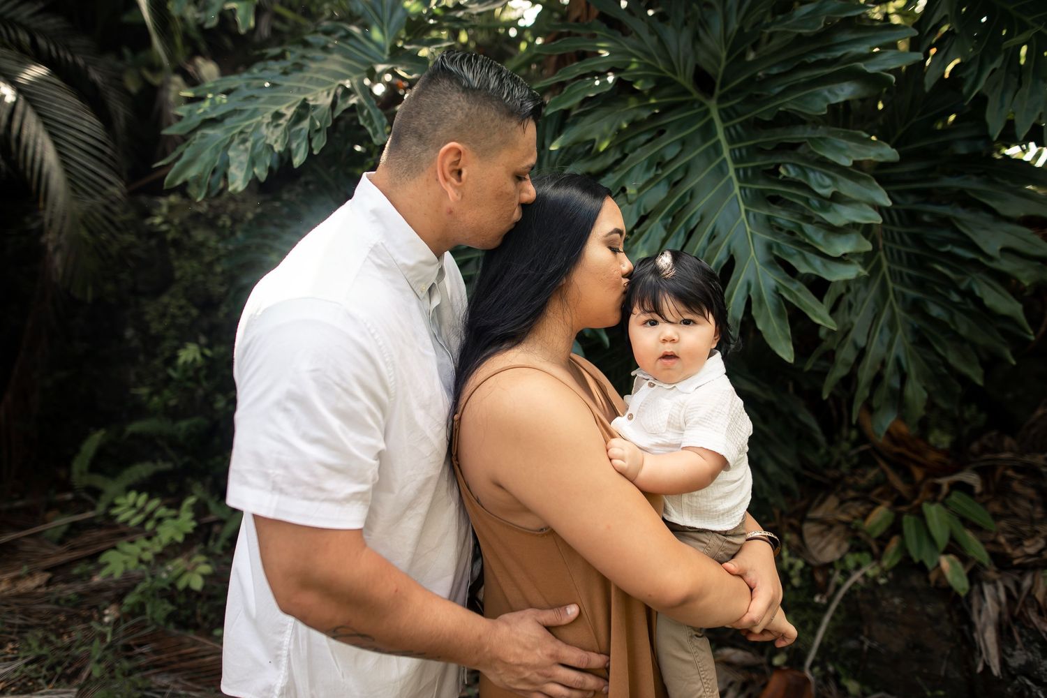 A family poses outdoors with lush tropical greenery, parents kissing their baby boy in a tender moment.