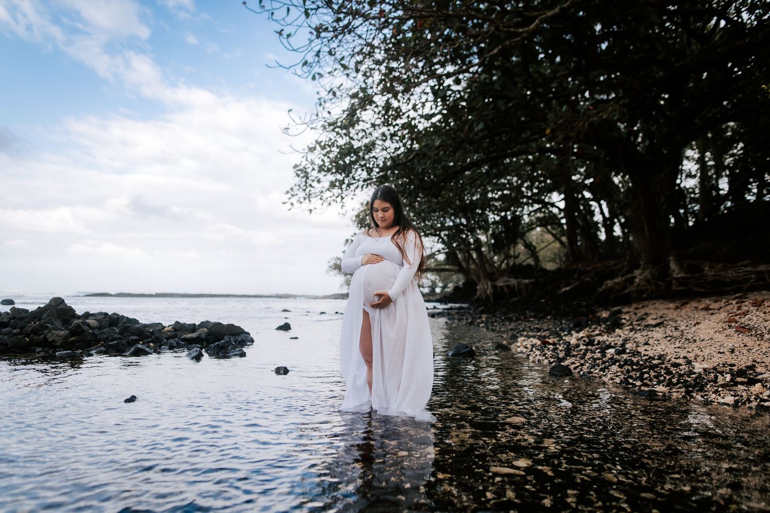 Pregnant woman in white flowing gown standing in shallow water near rocky shore with lush trees in background.