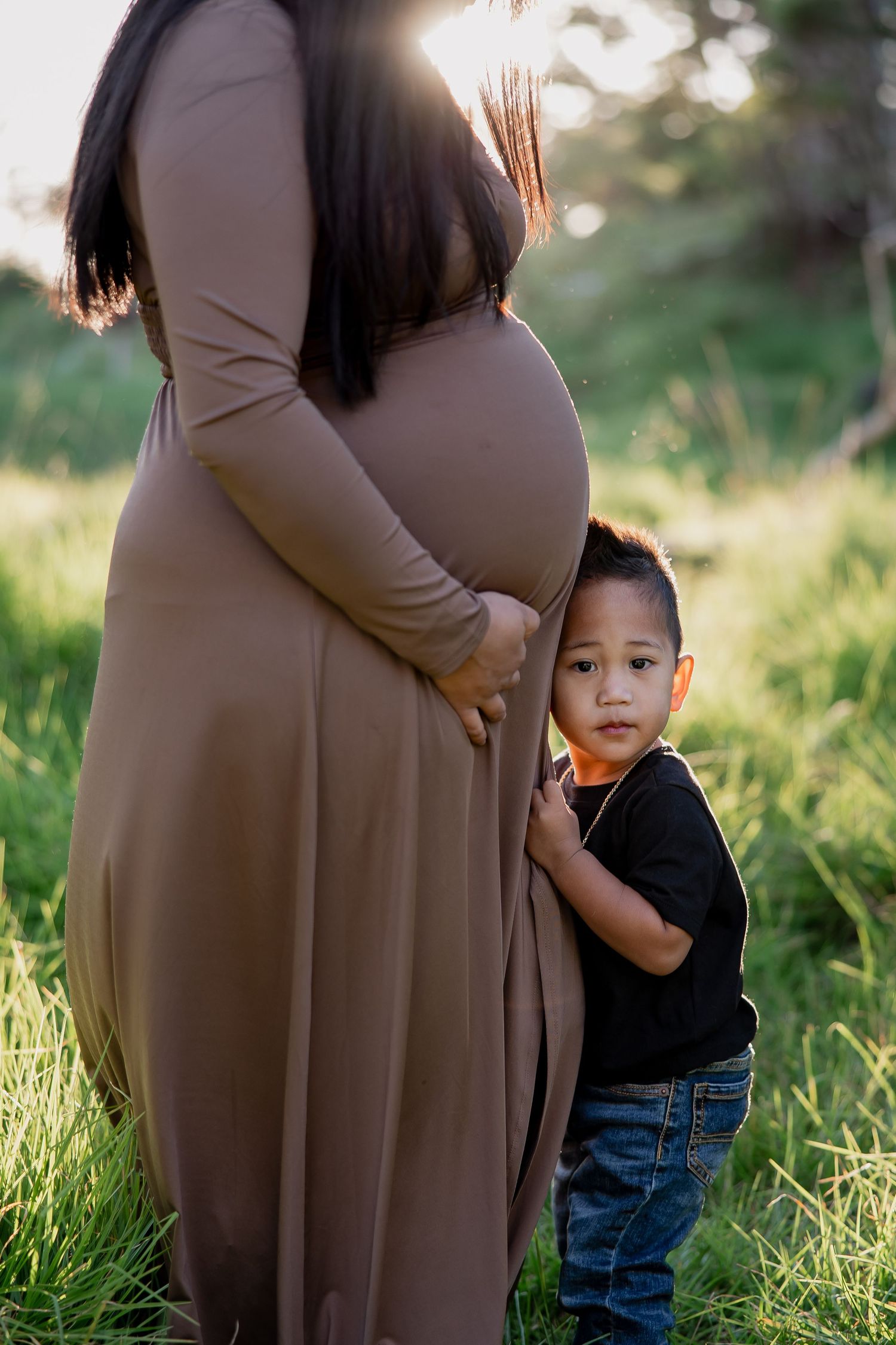 Pregnant woman in brown dress outdoors with young toddler boy hugging her belly in green grass field.