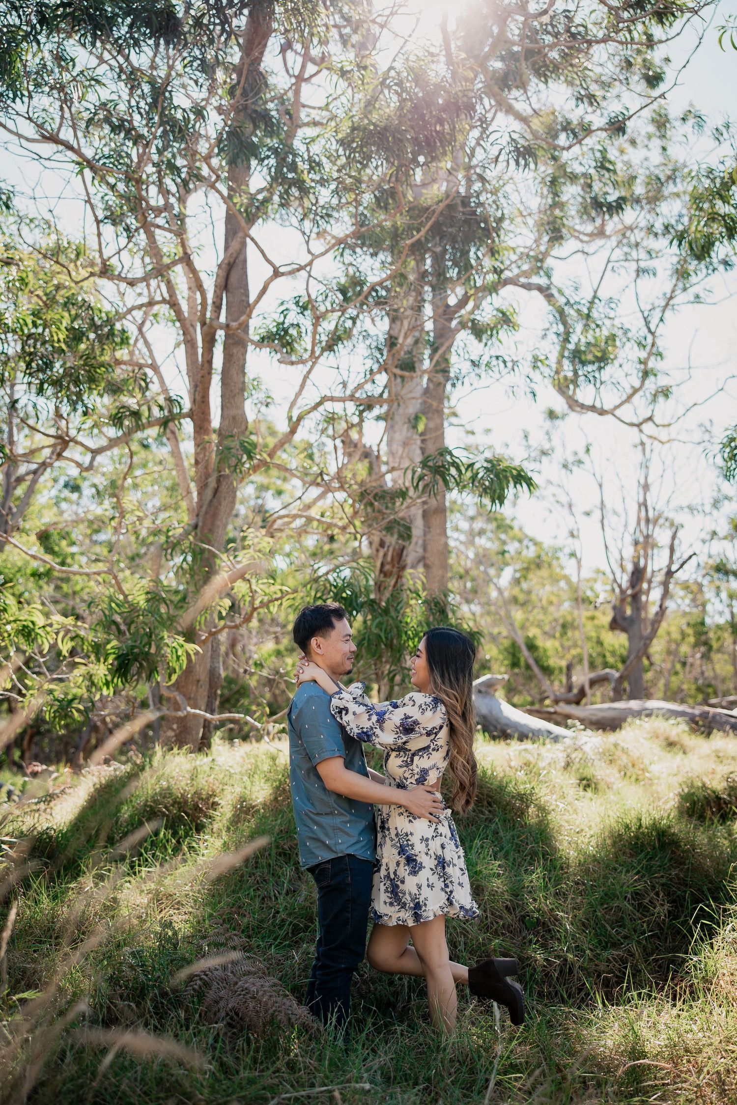 Couple embracing in a sunlit forest clearing, surrounded by tall eucalyptus trees and lush green grass.