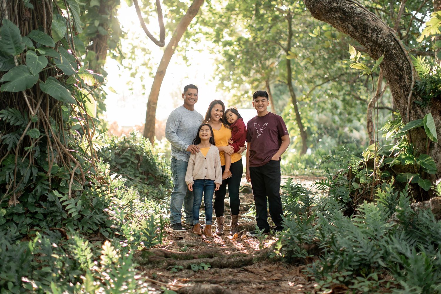Family of five posing together in a lush green forest pathway with sunlight filtering through the trees.