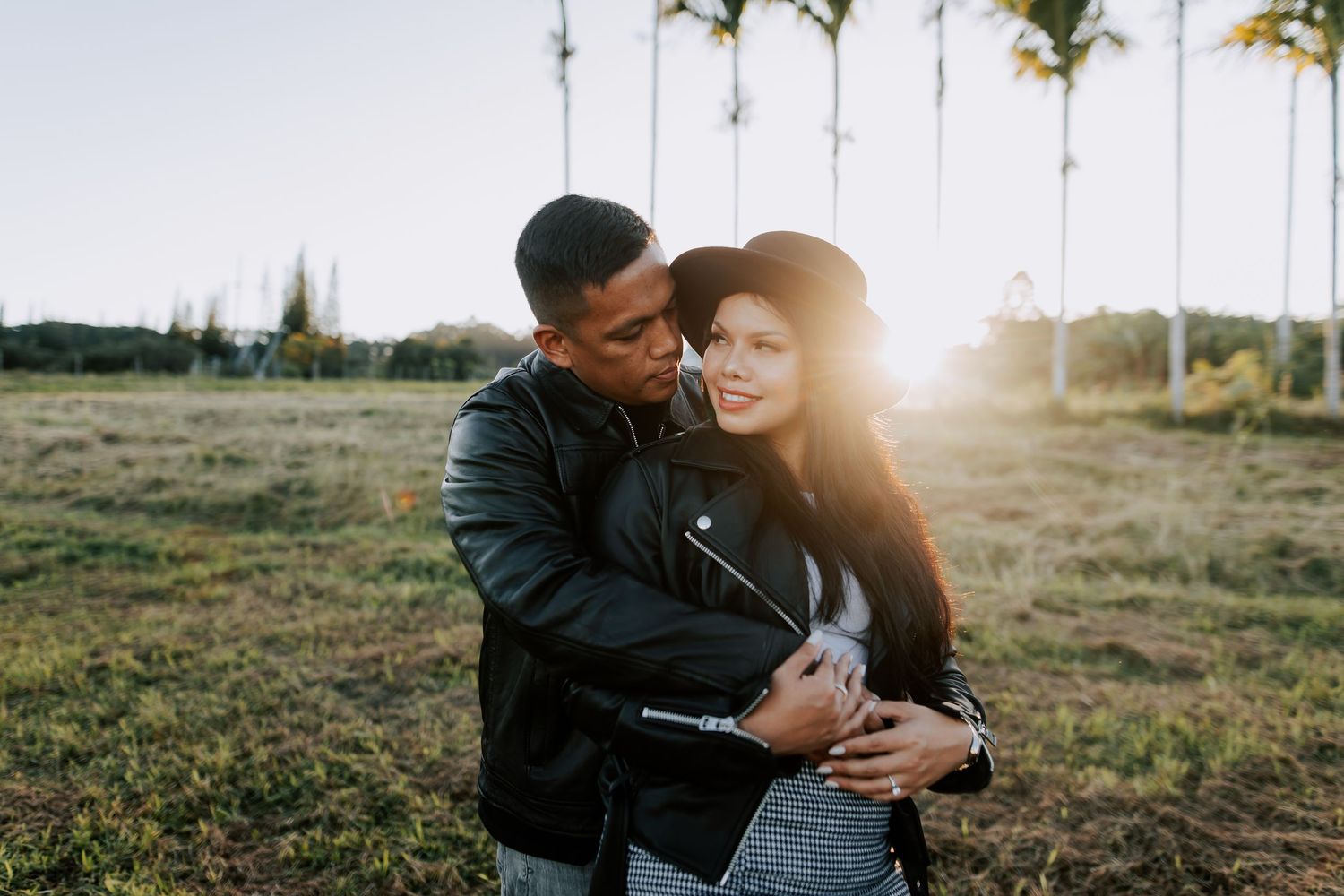 Couple embracing outdoors at golden hour, man in leather jacket hugging woman in black hat, palm trees in background.