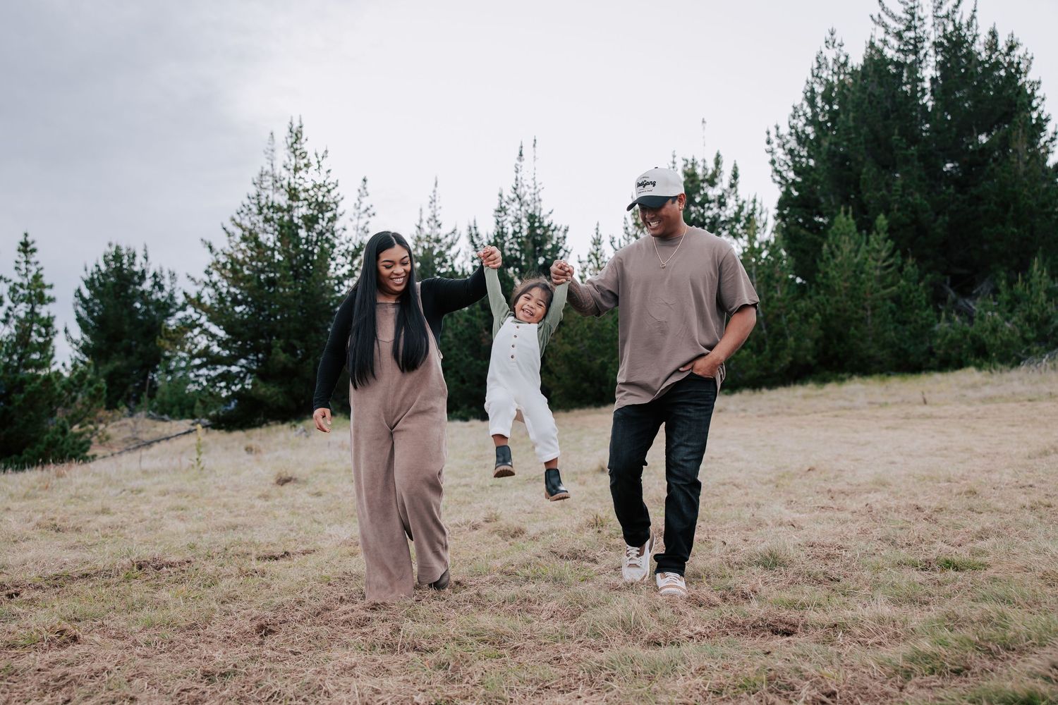 Family of three outdoors, parents swinging toddler between them in a grassy field with pine trees.