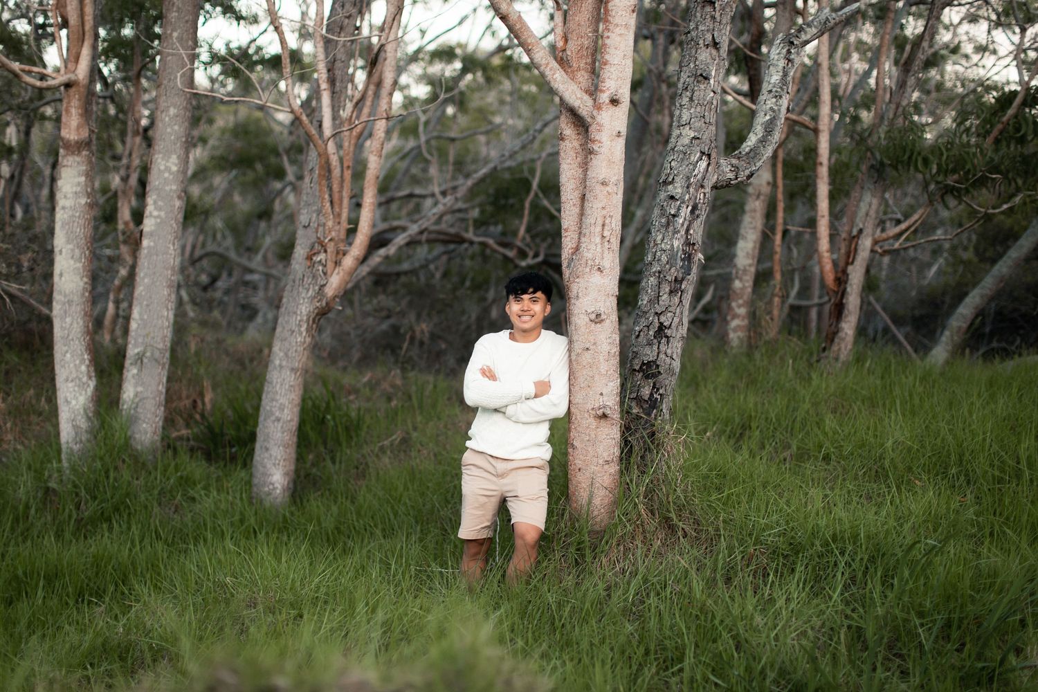 A young boy with crossed arms stands beside a white tree trunk in a lush green forest clearing.