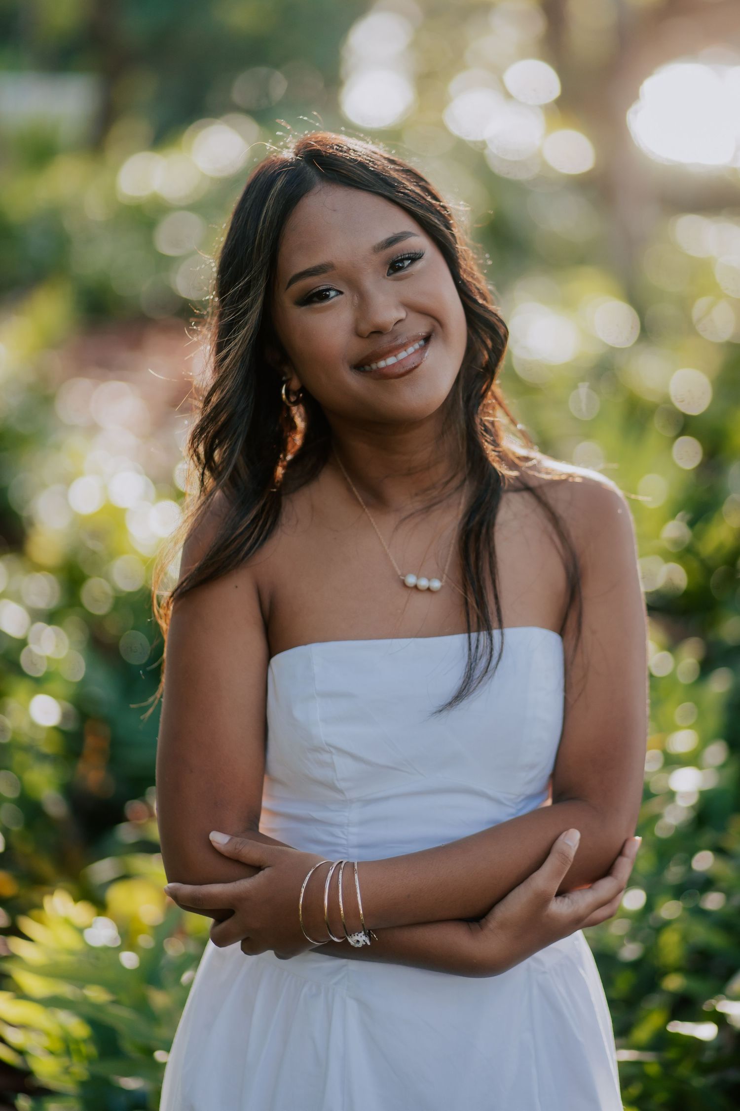 Young woman in white strapless dress smiling outdoors with lush green bokeh background.