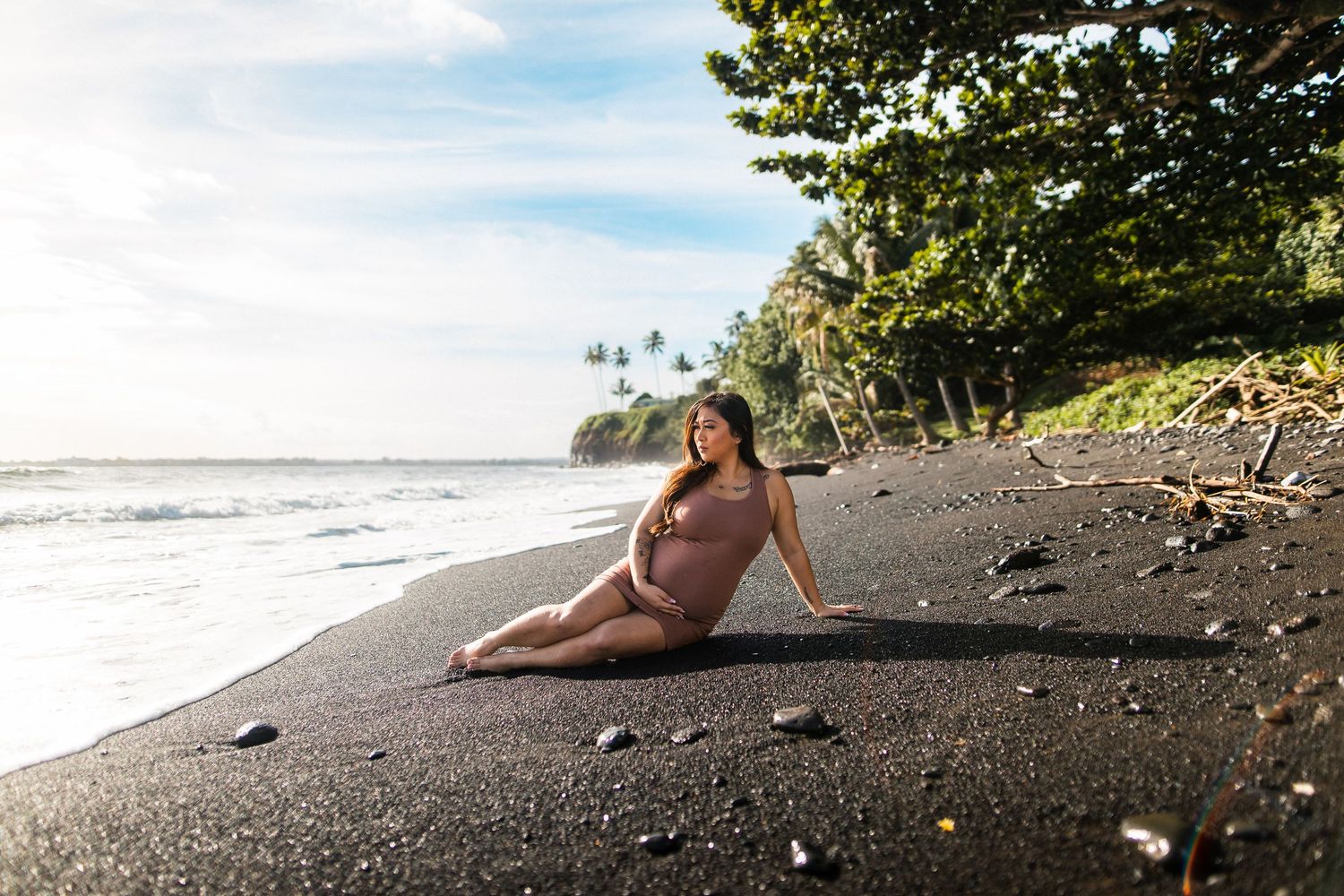 Pregnant woman in nude swimsuit posing on black sand beach with ocean waves and palm trees in background.