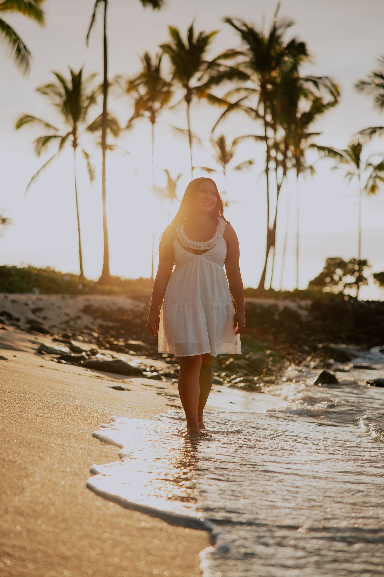 Woman in white dress walking barefoot on tropical beach at golden sunset with palm trees in background.