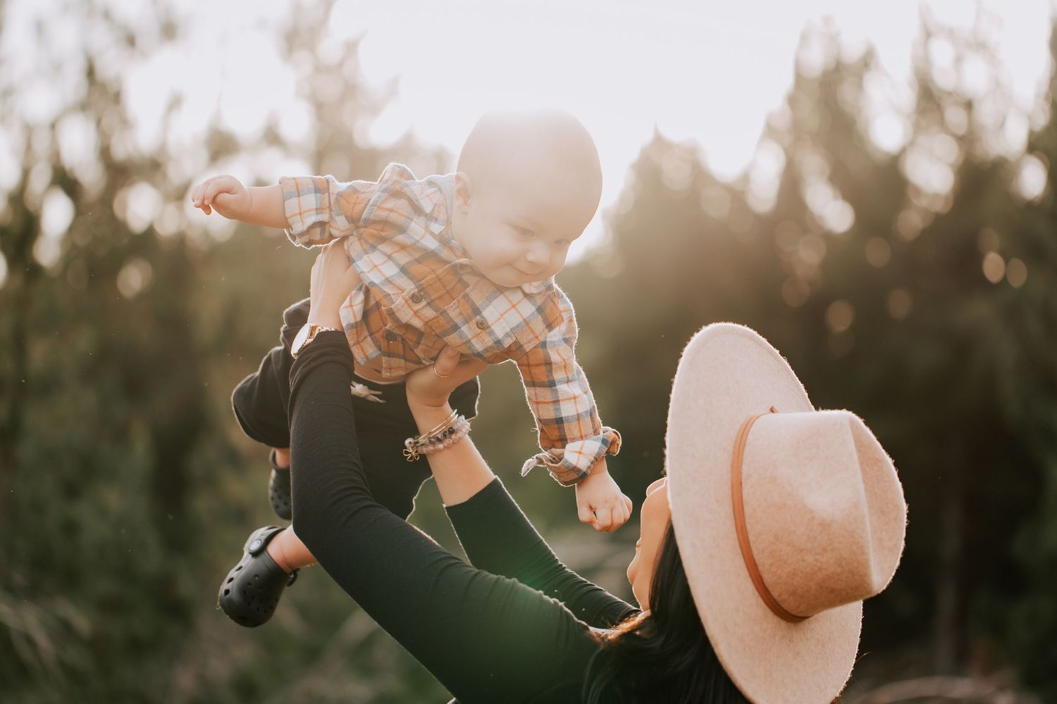 A mother in a sun hat lifts her smiling baby into the air outdoors in golden sunlight.