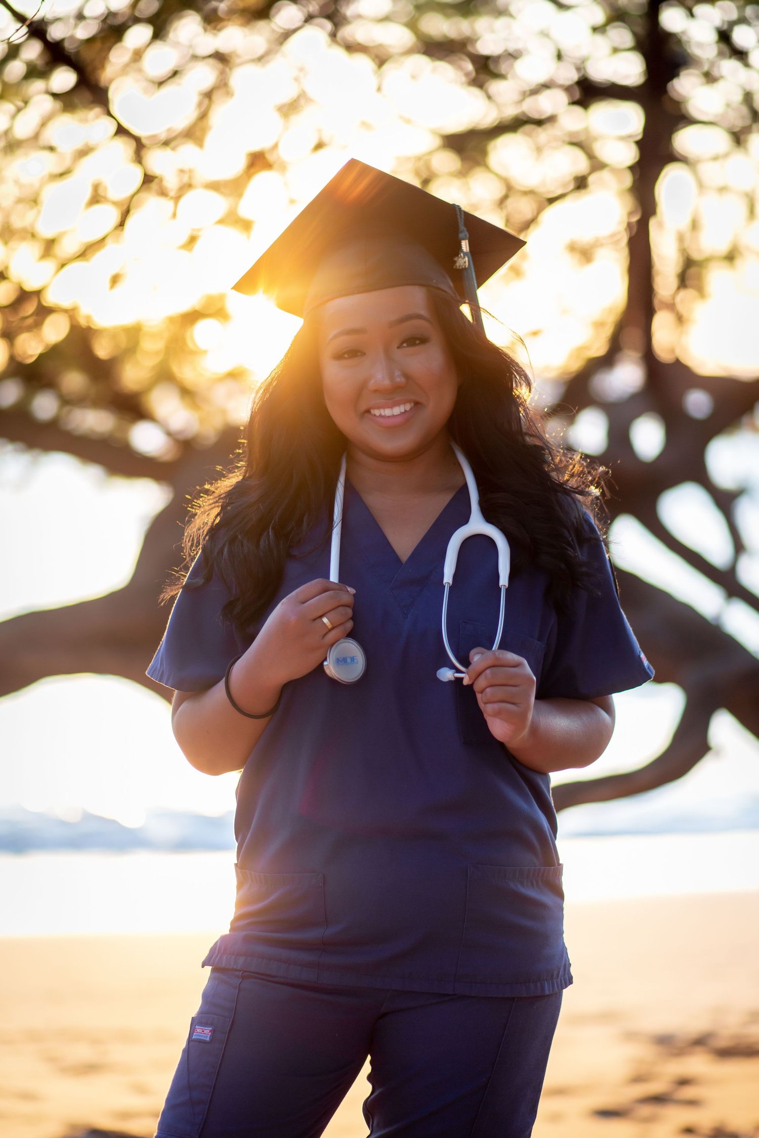 Smiling nursing graduate in blue scrubs and graduation cap with stethoscope, posing outdoors at sunset near a tree.