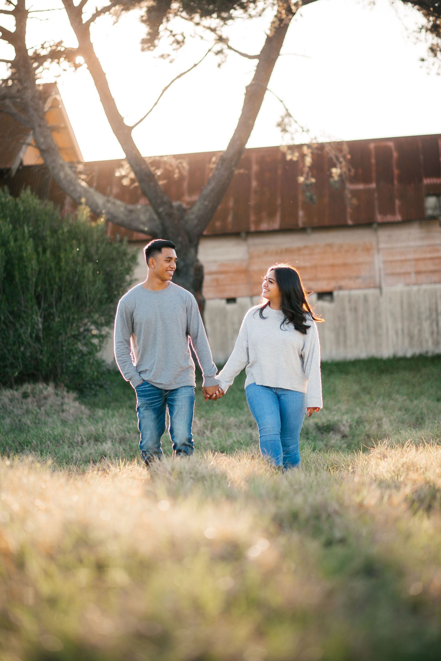 Couple holding hands walking through golden sunlit field near rustic barn, wearing casual jeans and sweaters.
