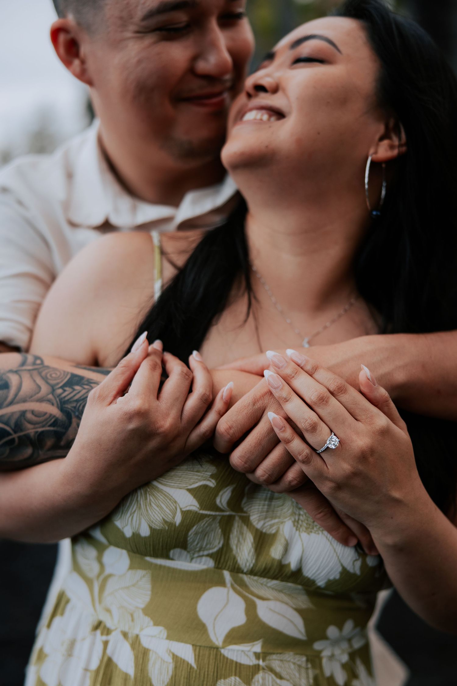 Smiling couple embracing outdoors, woman wearing engagement ring and floral dress, man with tattoo hugging her from behind.
