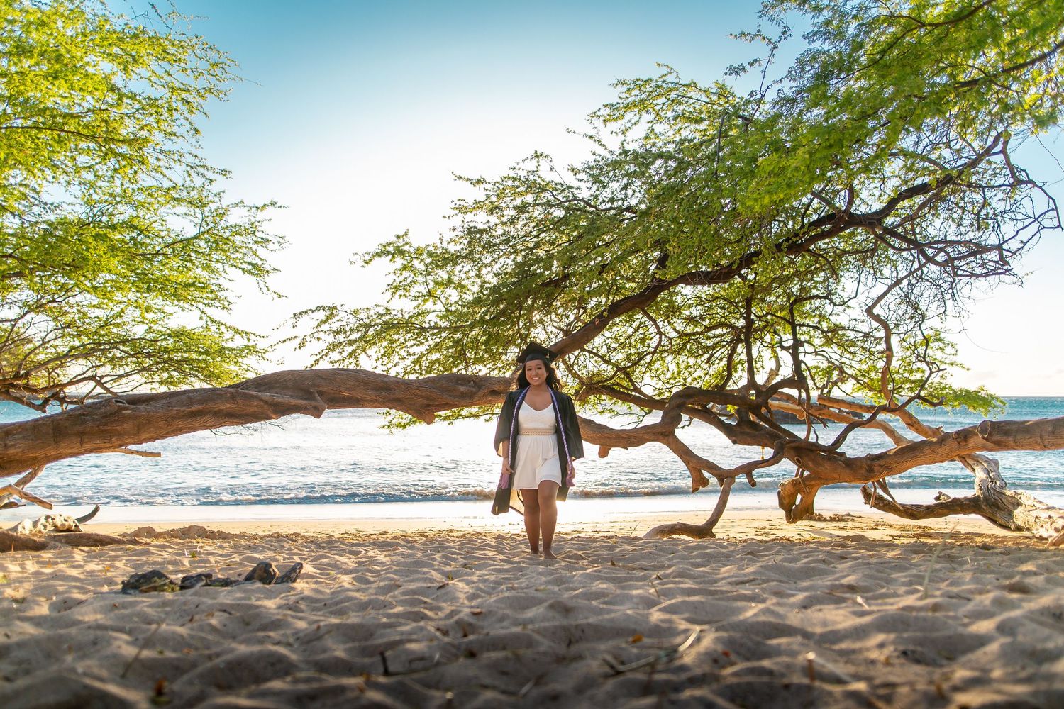 Woman walking on sandy beach under sprawling tree branches with ocean and blue sky in background.