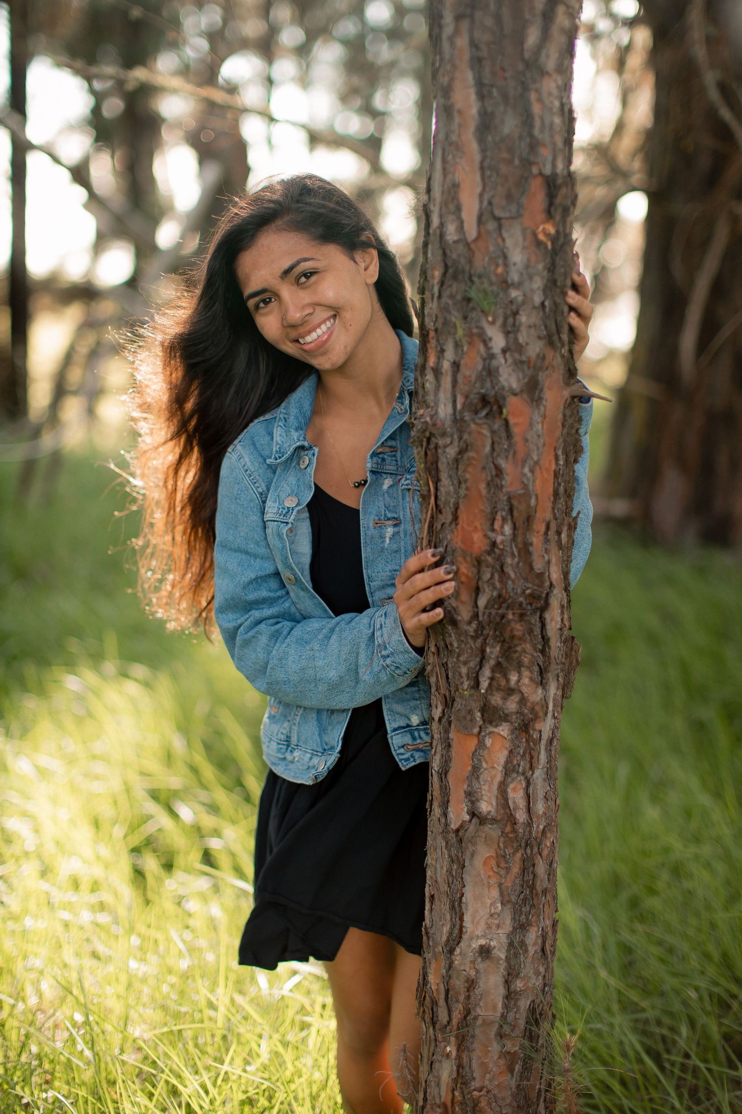 Young woman in denim jacket and black dress smiling beside a tree trunk in a sunlit forest.