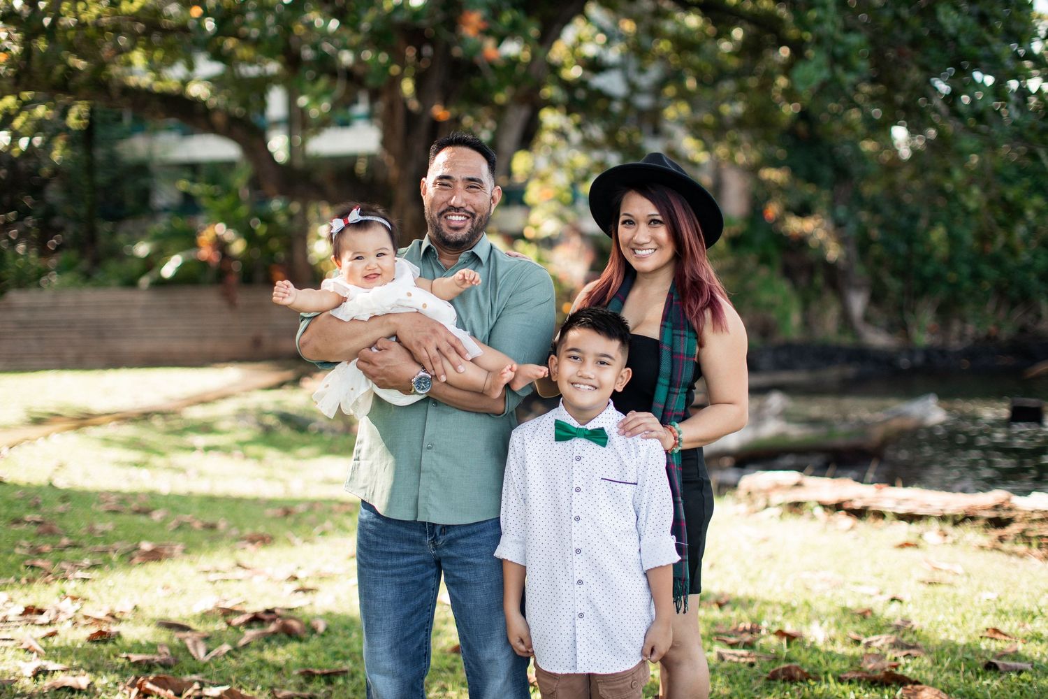 Happy family of four posing outdoors, parents holding baby and young boy in bow tie, smiling in a park setting.