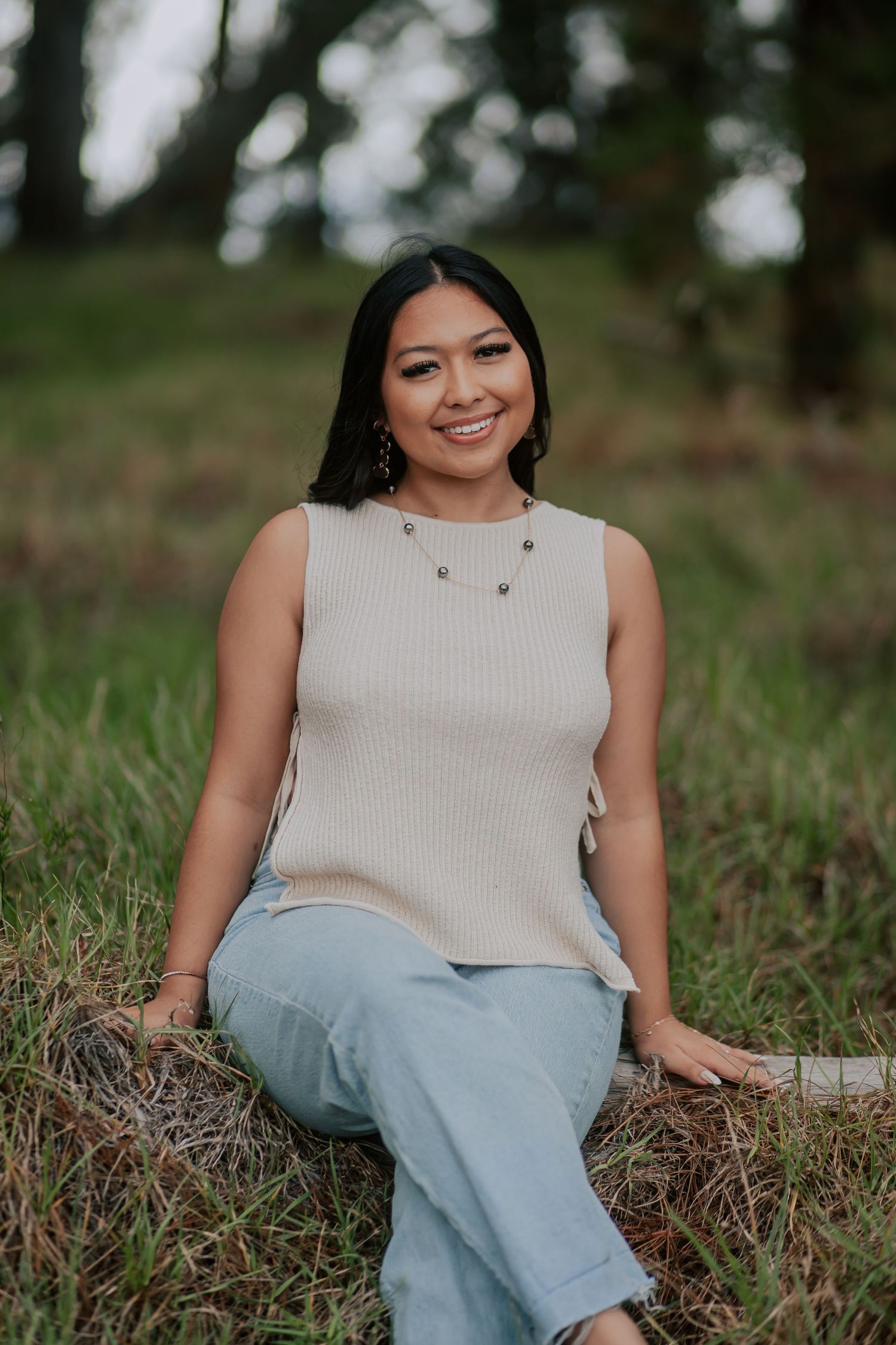 Young woman in beige ribbed top and light blue jeans smiling outdoors, seated on hay in a grassy field.
