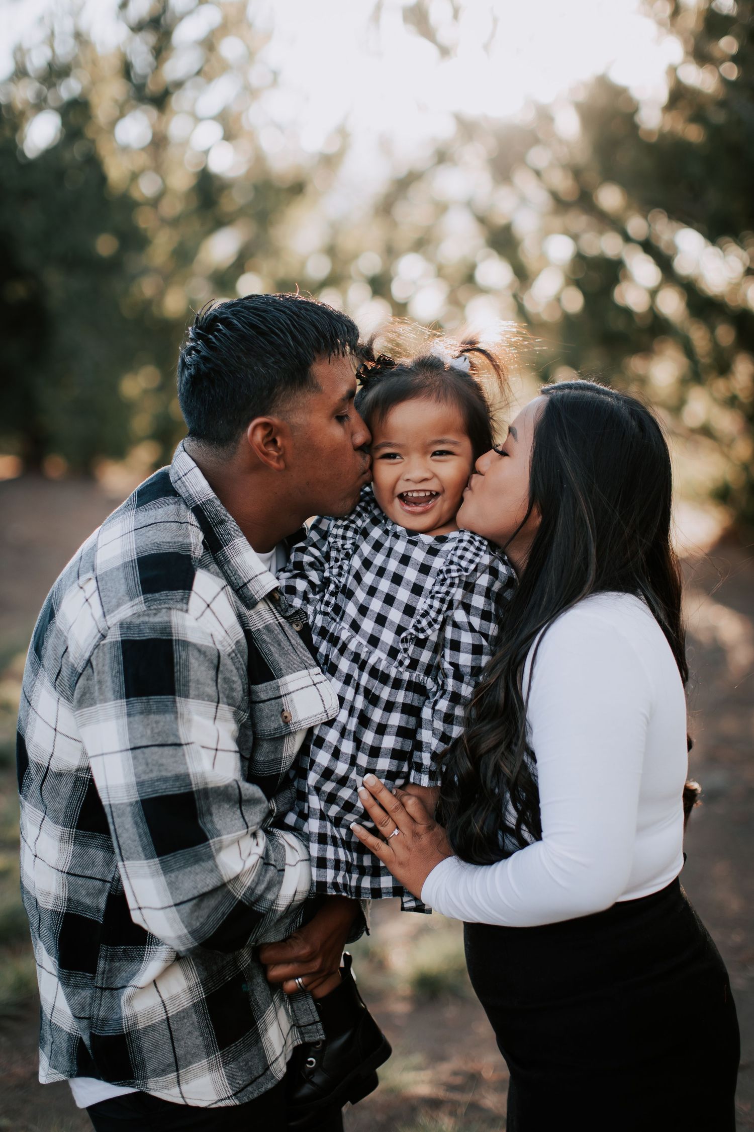 A joyful family portrait with parents kissing their laughing toddler daughter outdoors among trees.