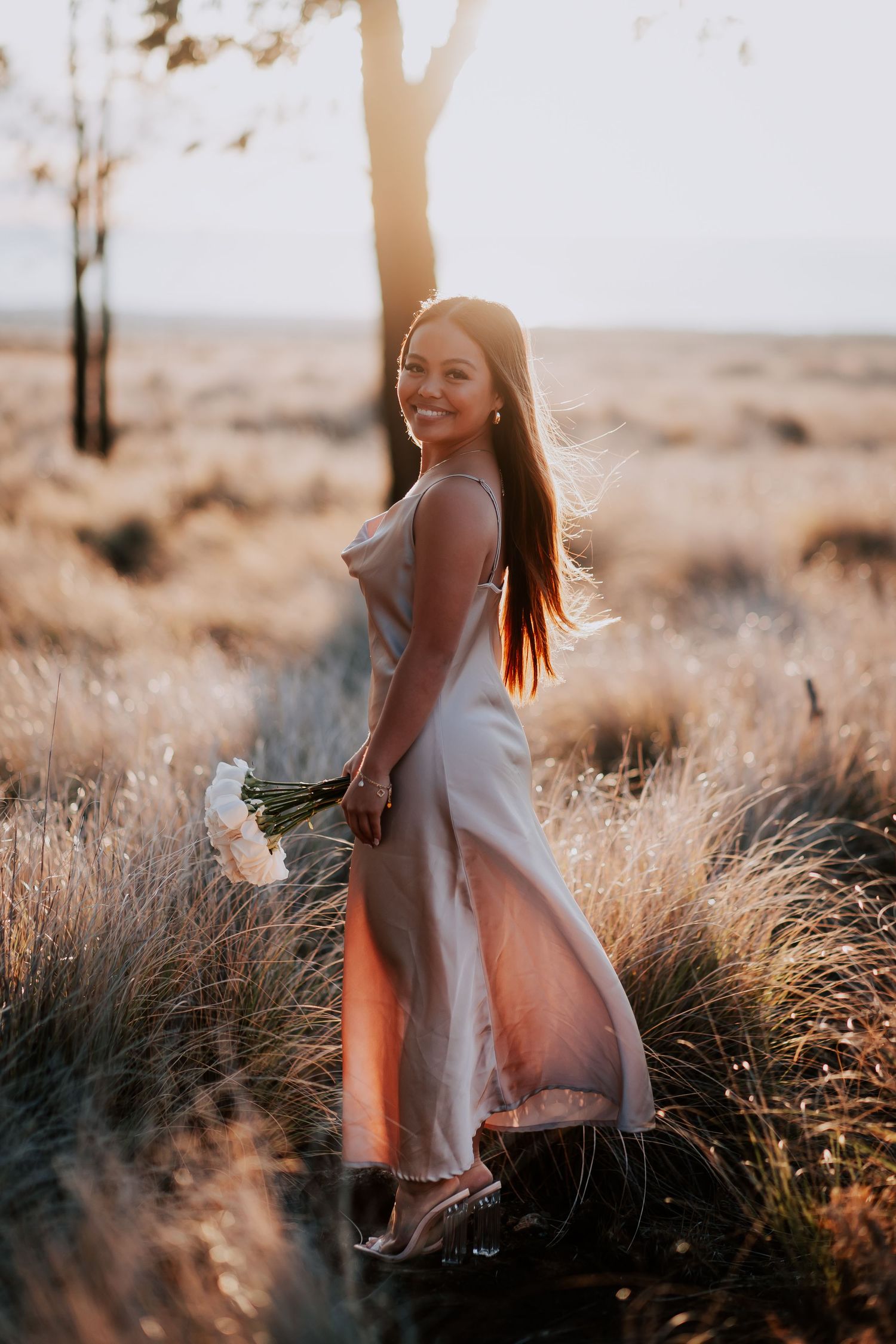 Woman in elegant satin dress holding white flowers, standing in golden field at sunset.