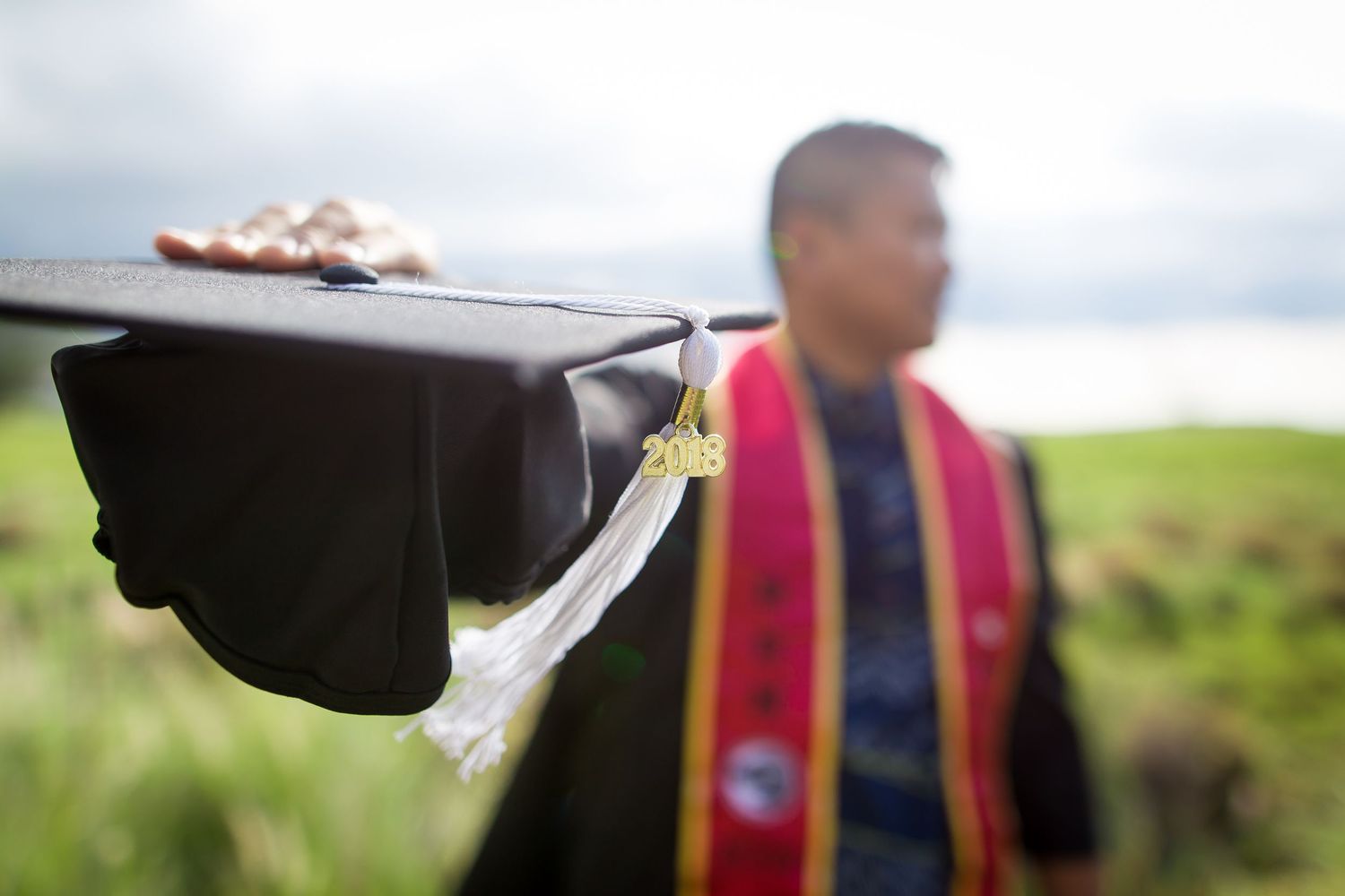 Graduate holding mortarboard cap outdoors wearing colorful sash, celebrating academic achievement in a scenic green field.