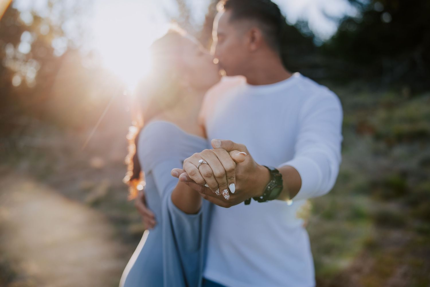 Couple kissing at sunset while displaying engagement ring, golden backlight illuminating romantic outdoor proposal moment.