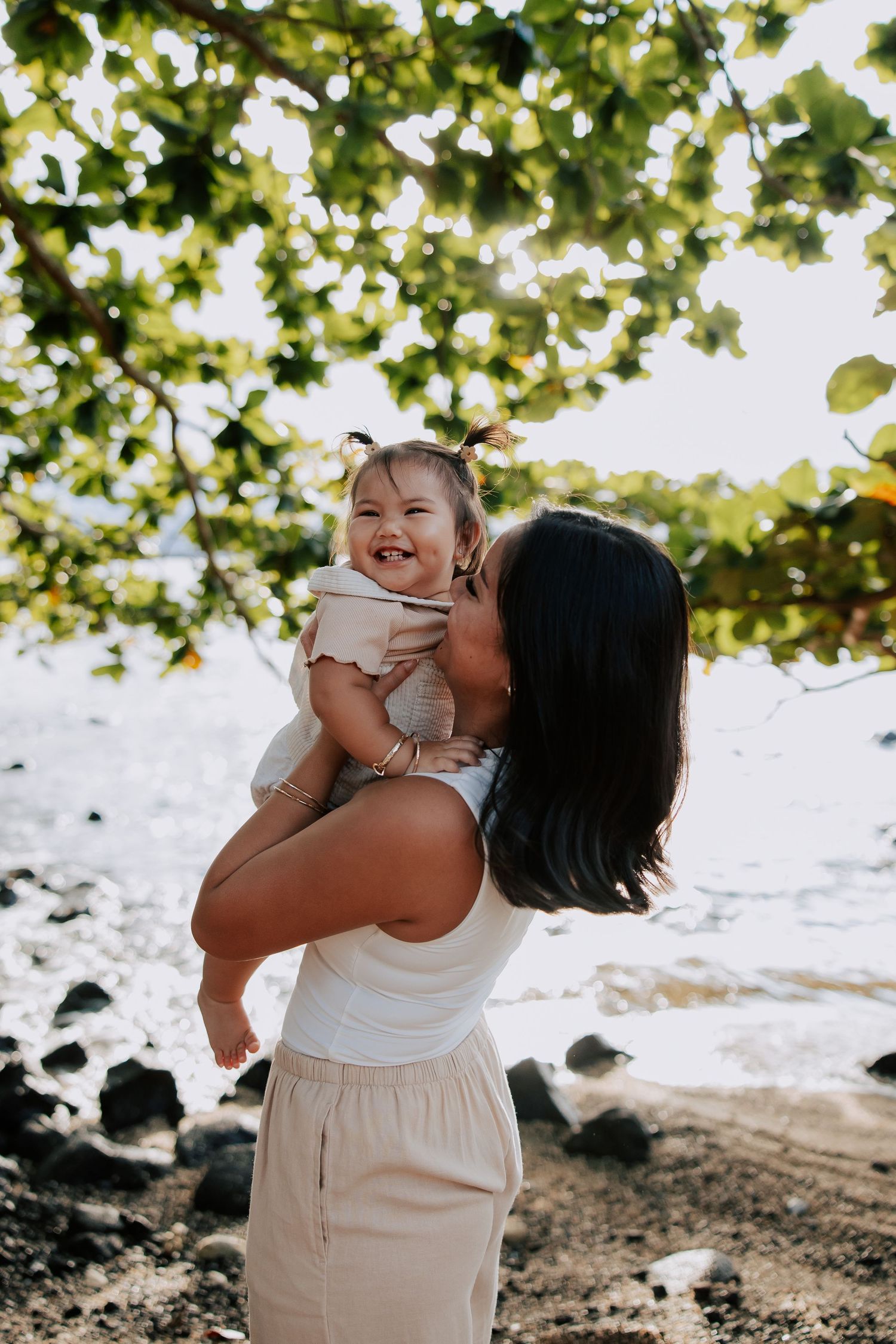 A mother kisses her laughing baby girl on a rocky beach under green trees.