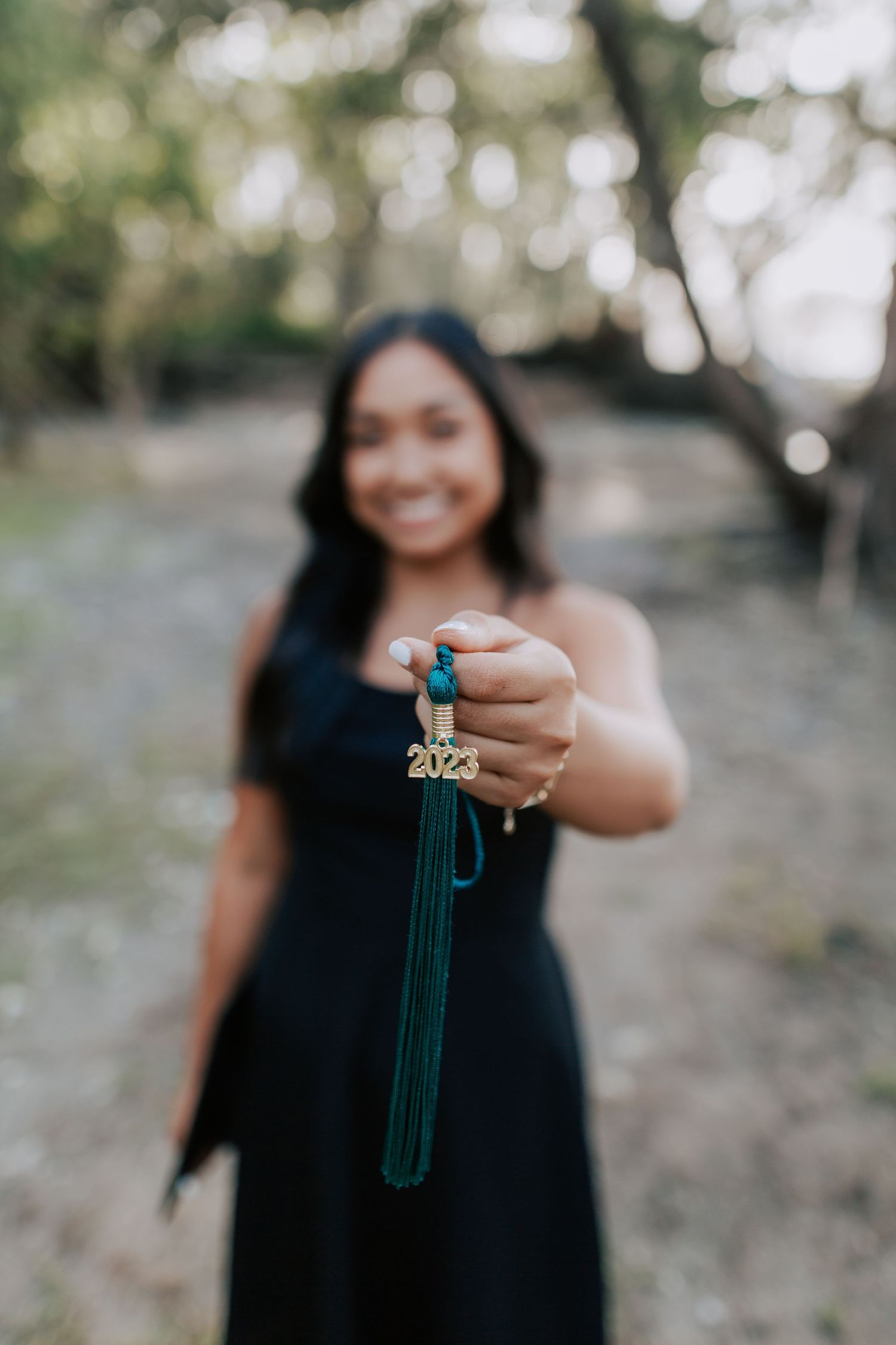 Smiling graduate in black dress holds green 2023 tassel toward camera outdoors.
