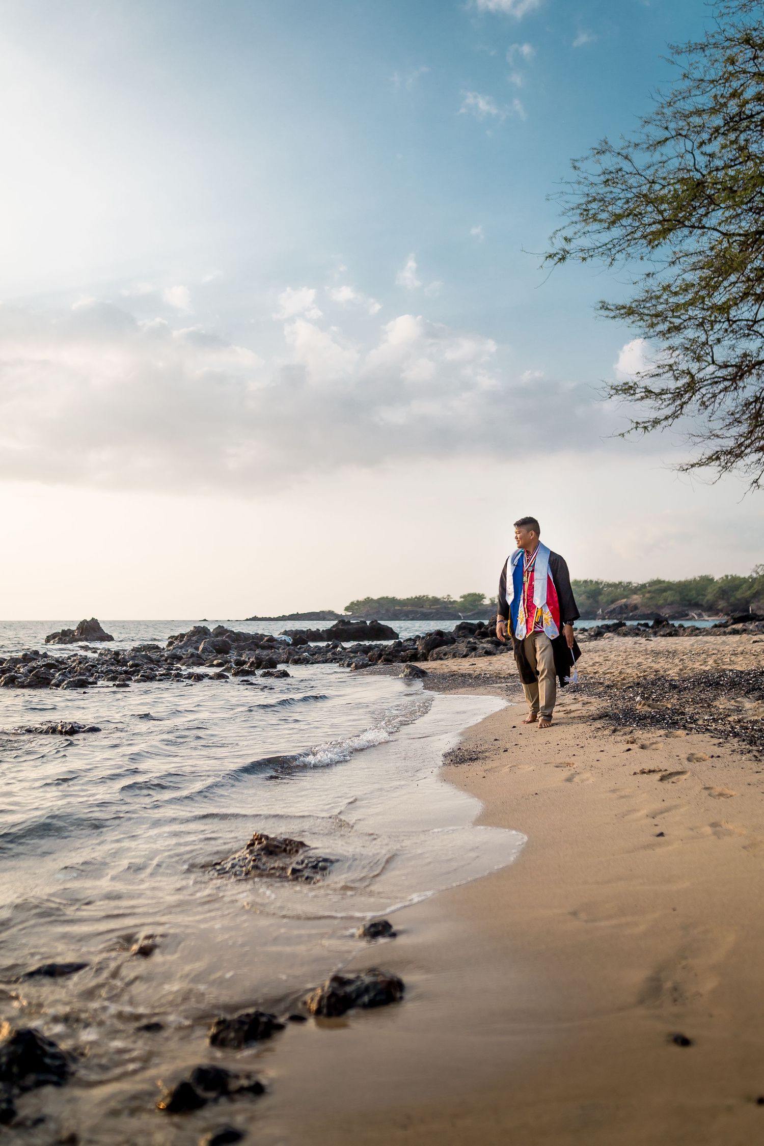 A man wrapped in a colorful towel walks barefoot along a rocky Hawaiian beach at sunset.