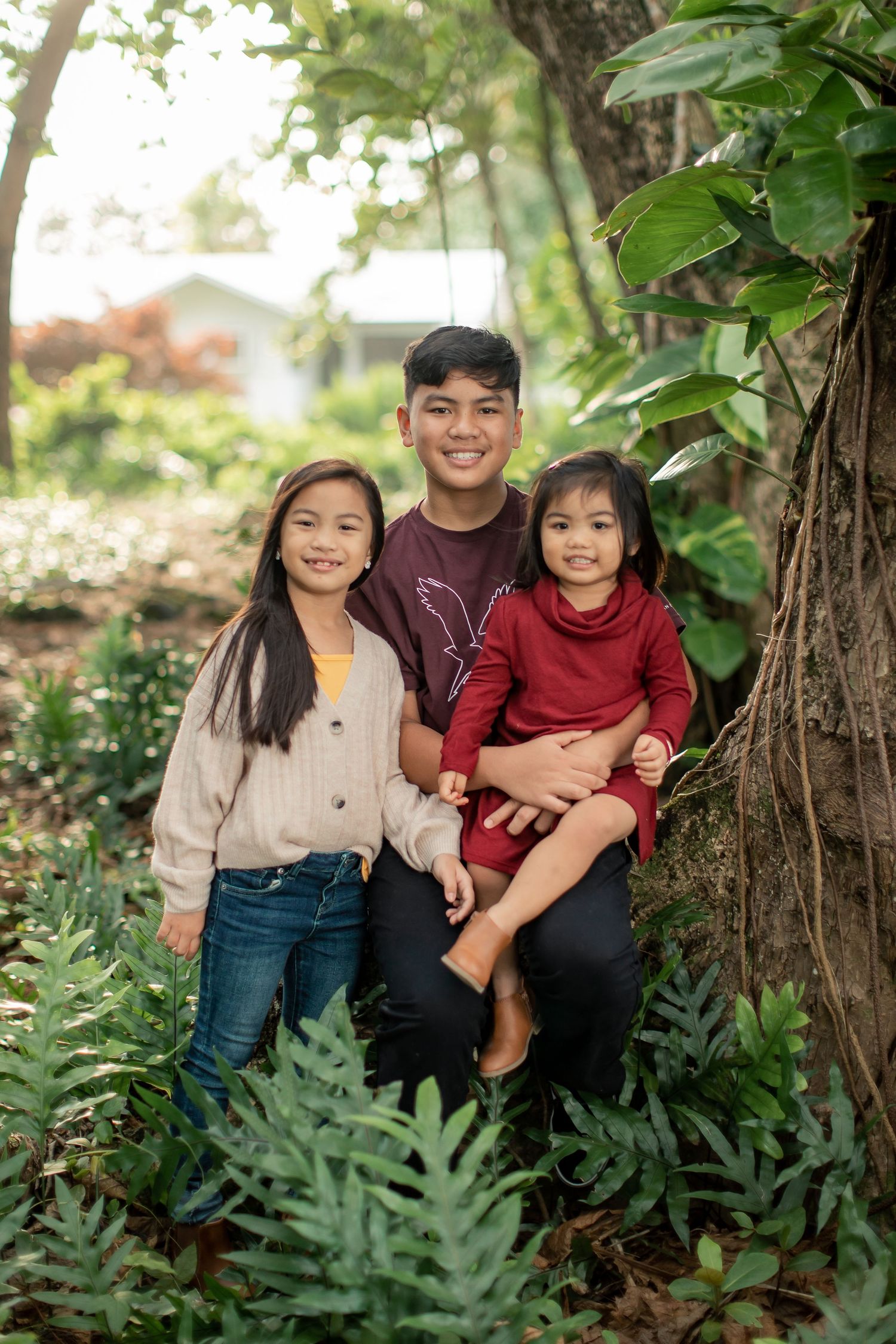 A young man smiles with two girls beside a tree in a lush garden, posing for a cheerful family portrait outdoors.