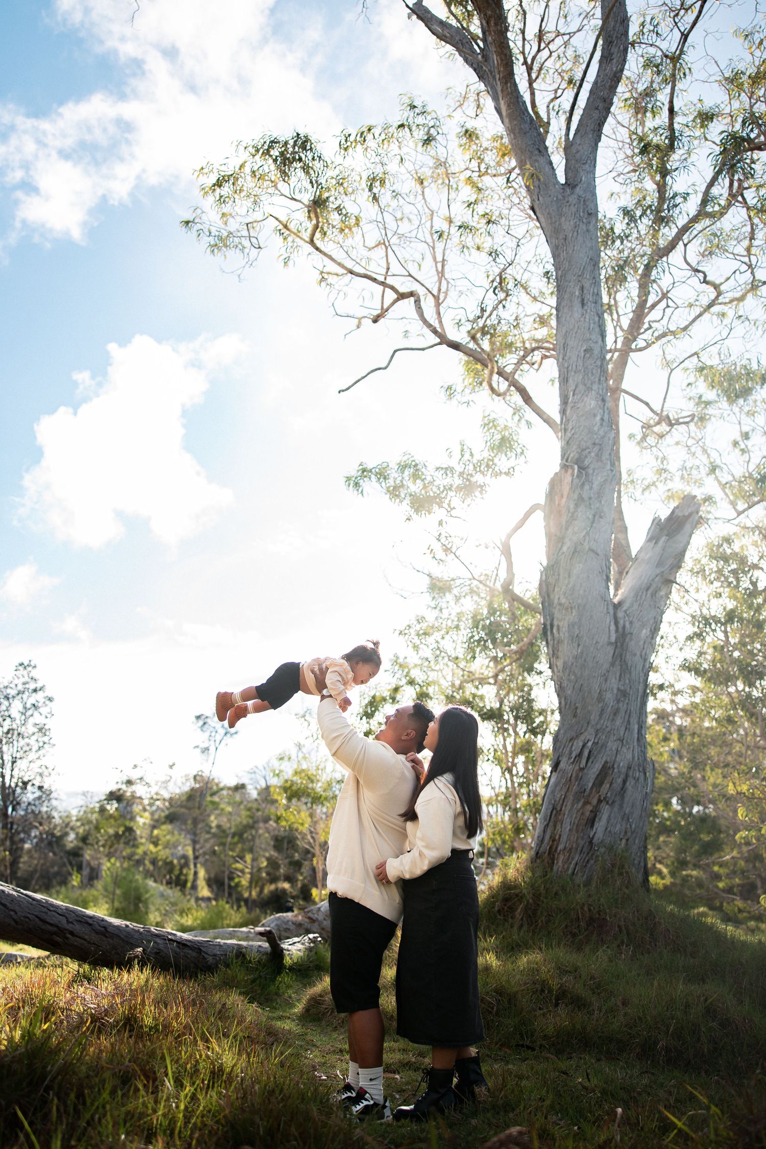 A couple lifts their baby joyfully in a sunlit park beside a tall eucalyptus tree.