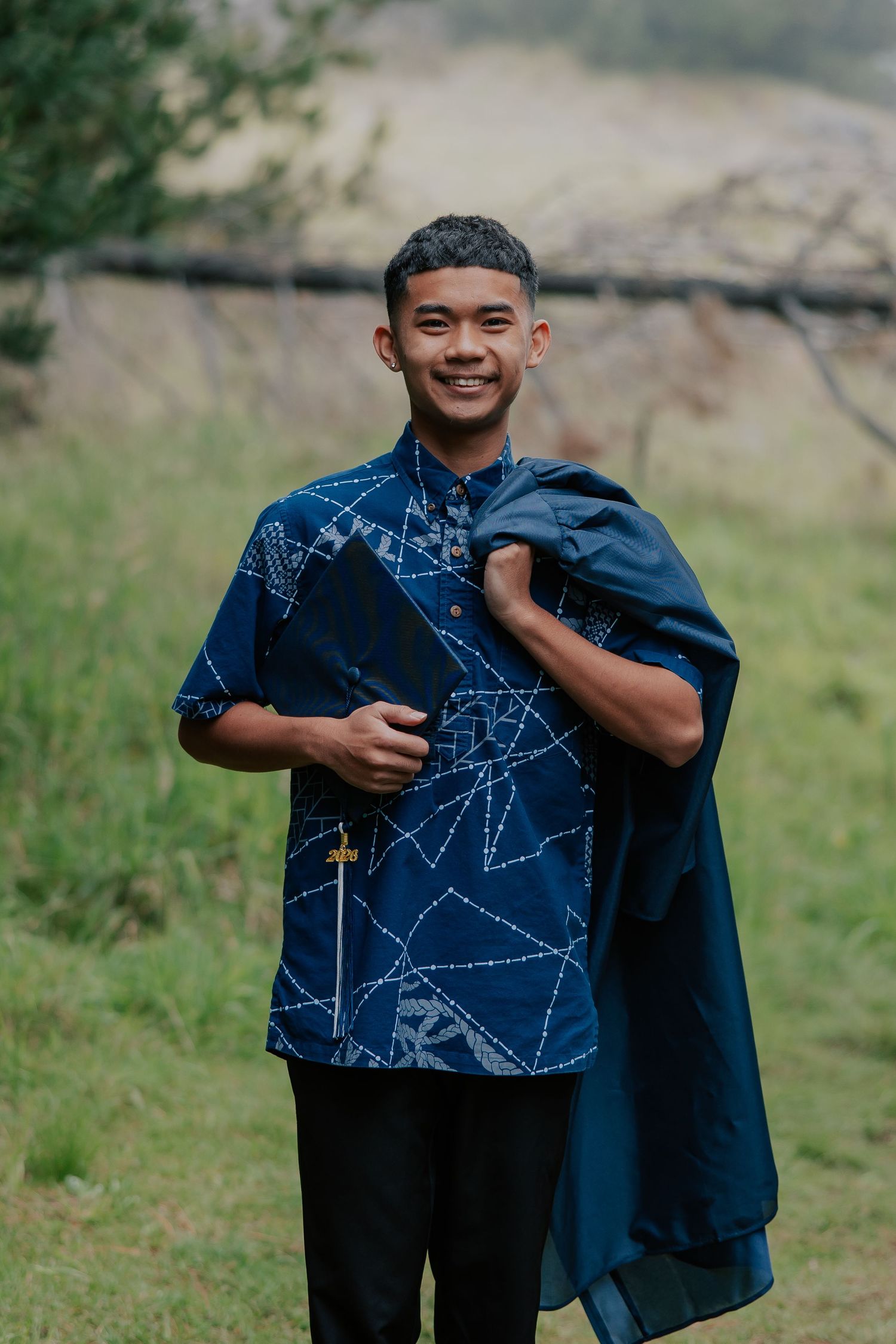 Young man smiling outdoors in a navy blue geometric print batik shirt, holding a jacket over his shoulder.