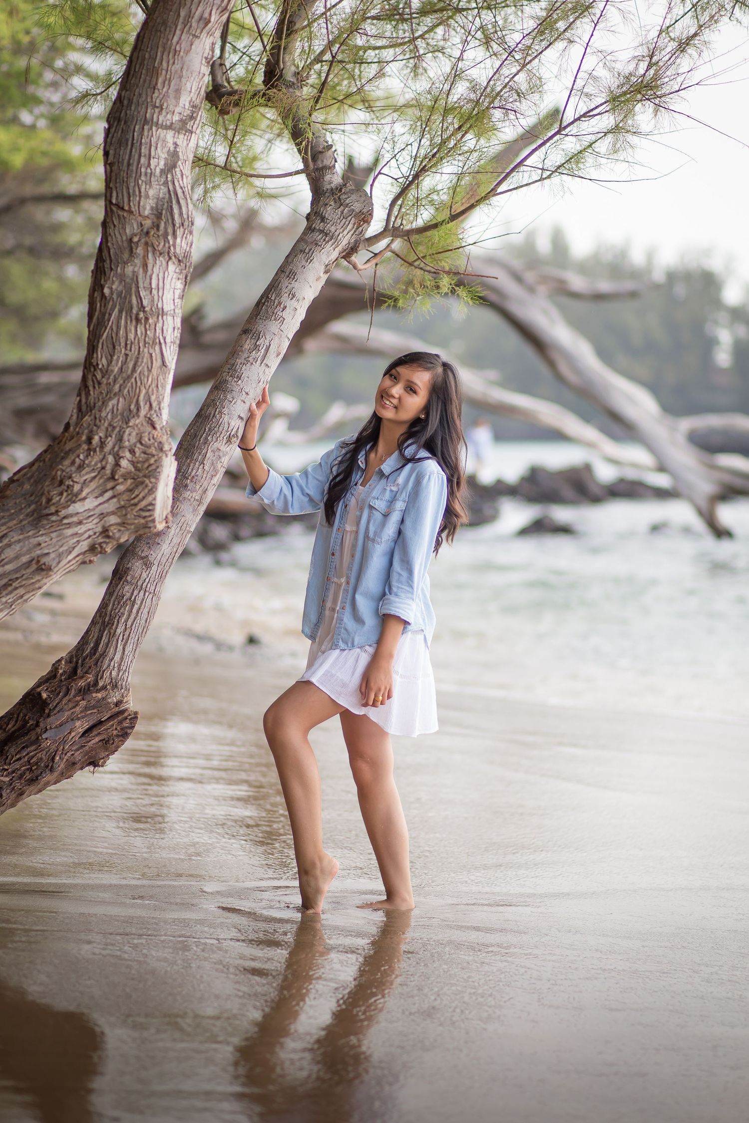 Young woman in denim shirt and white skirt standing barefoot on a tropical beach, leaning against a tree.