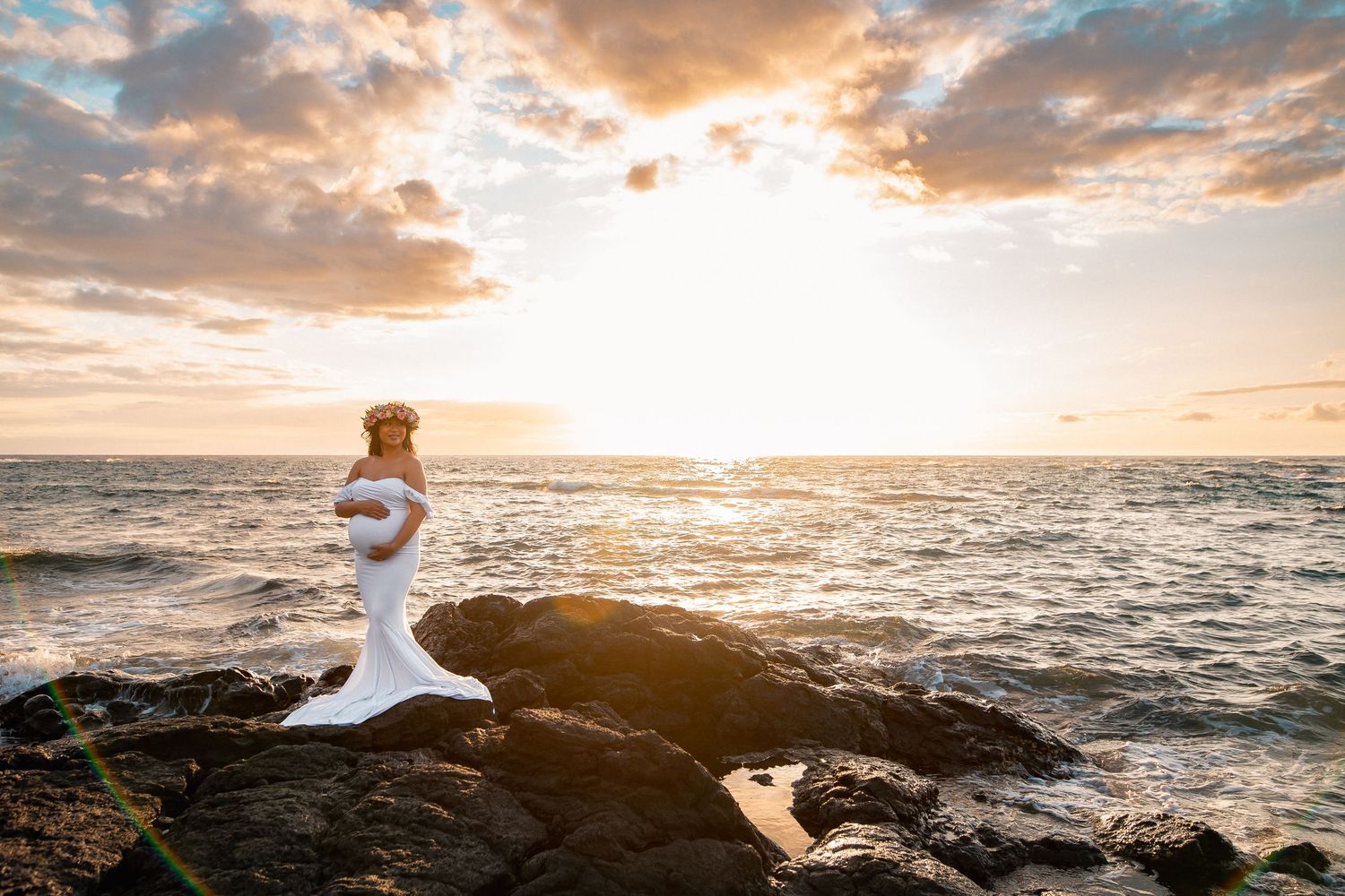 Pregnant woman in white gown poses on rocky shore at sunset with ocean backdrop in Hawaii.