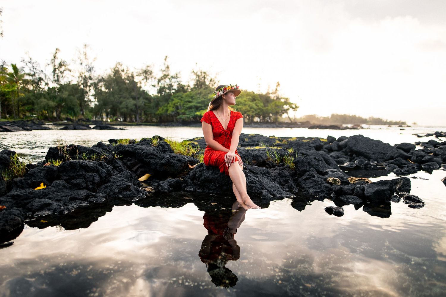 Woman in red dress sitting on volcanic rocks by a tropical shoreline with calm reflective water and lush trees.