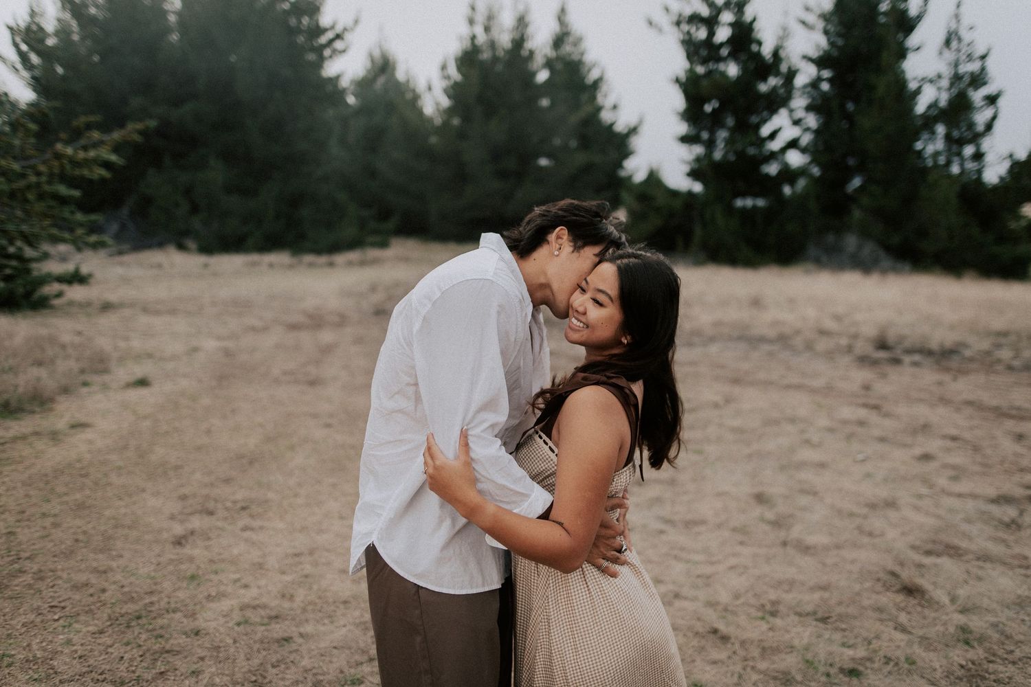 Couple embracing in a golden field surrounded by pine trees, man kissing woman's head in a romantic outdoor engagement photo.