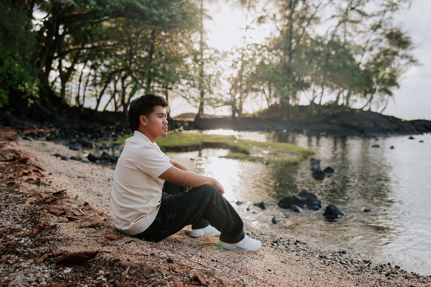 Young man sitting on rocky shoreline gazing at calm water surrounded by lush tropical trees at golden hour.