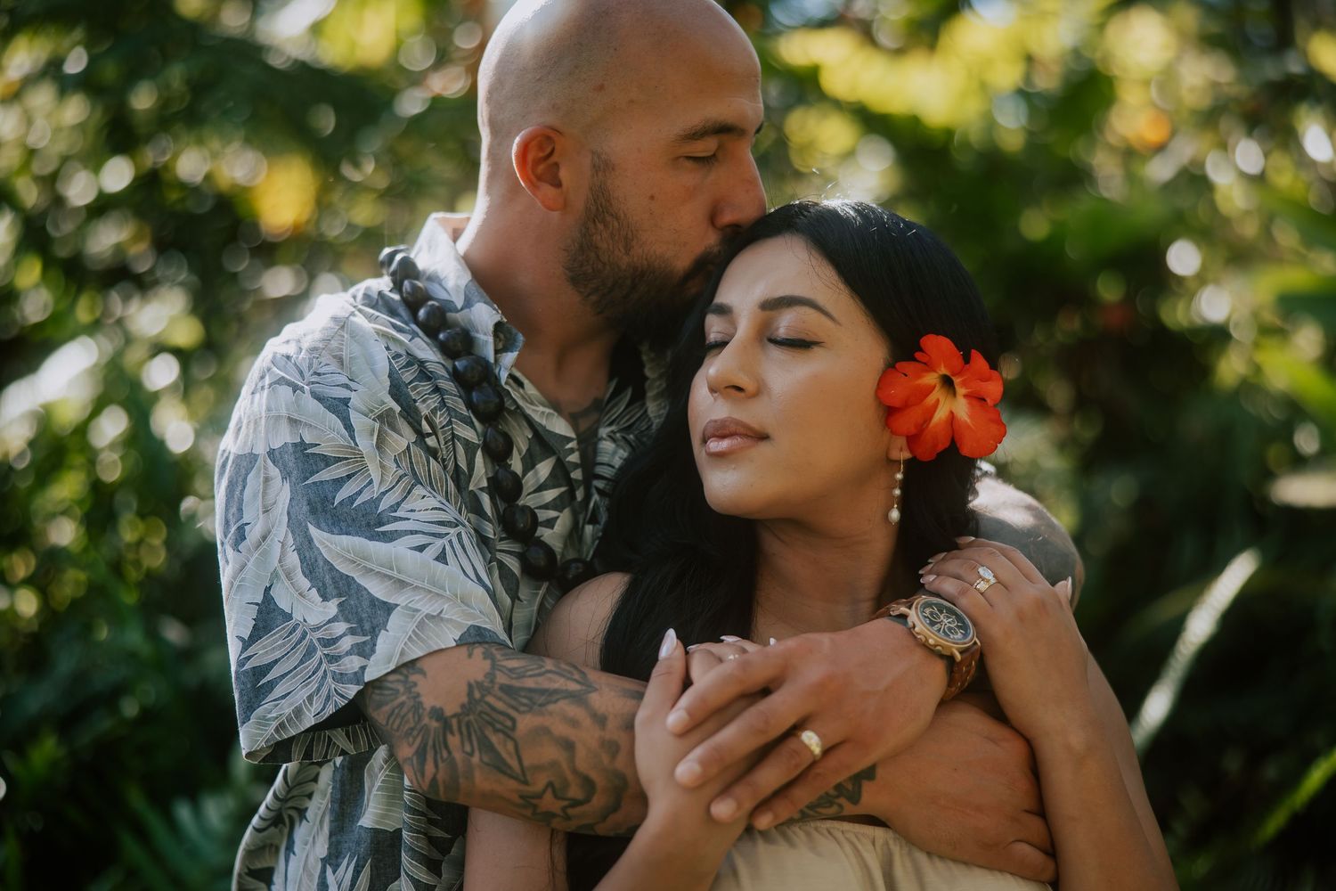 A couple embraces in a tropical garden, man kissing womans head, she wears an orange flower in her hair.