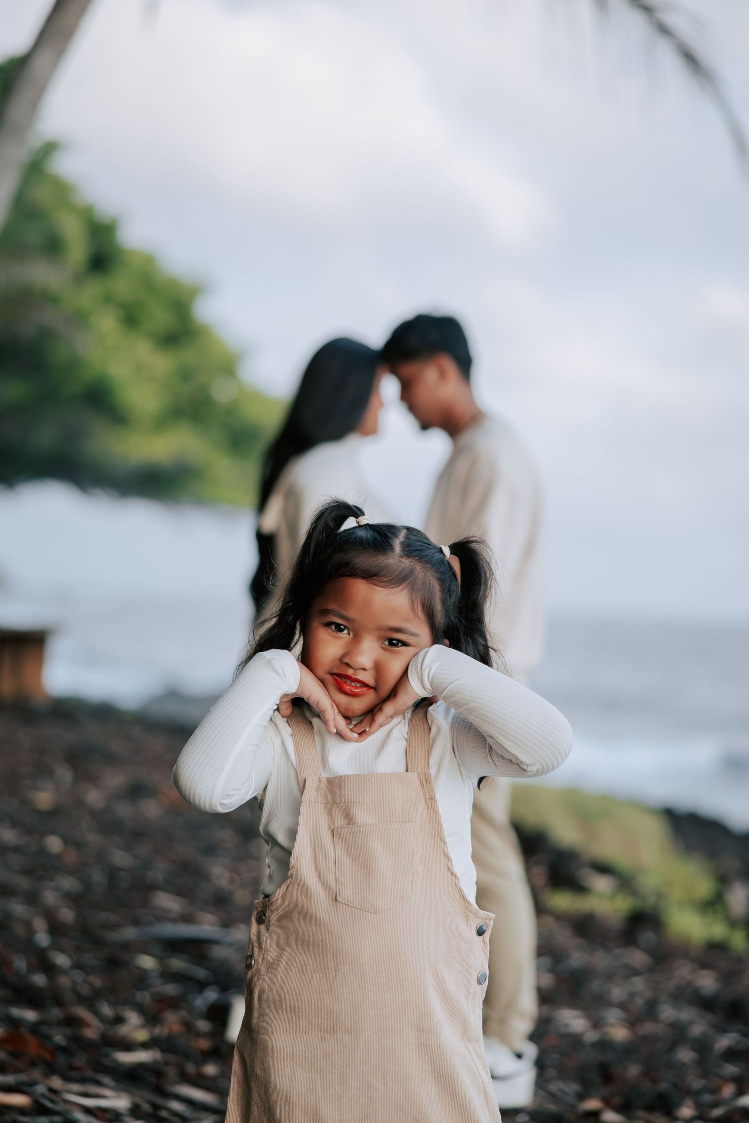 Adorable little girl in overalls poses on rocky beach while parents share romantic moment in background.