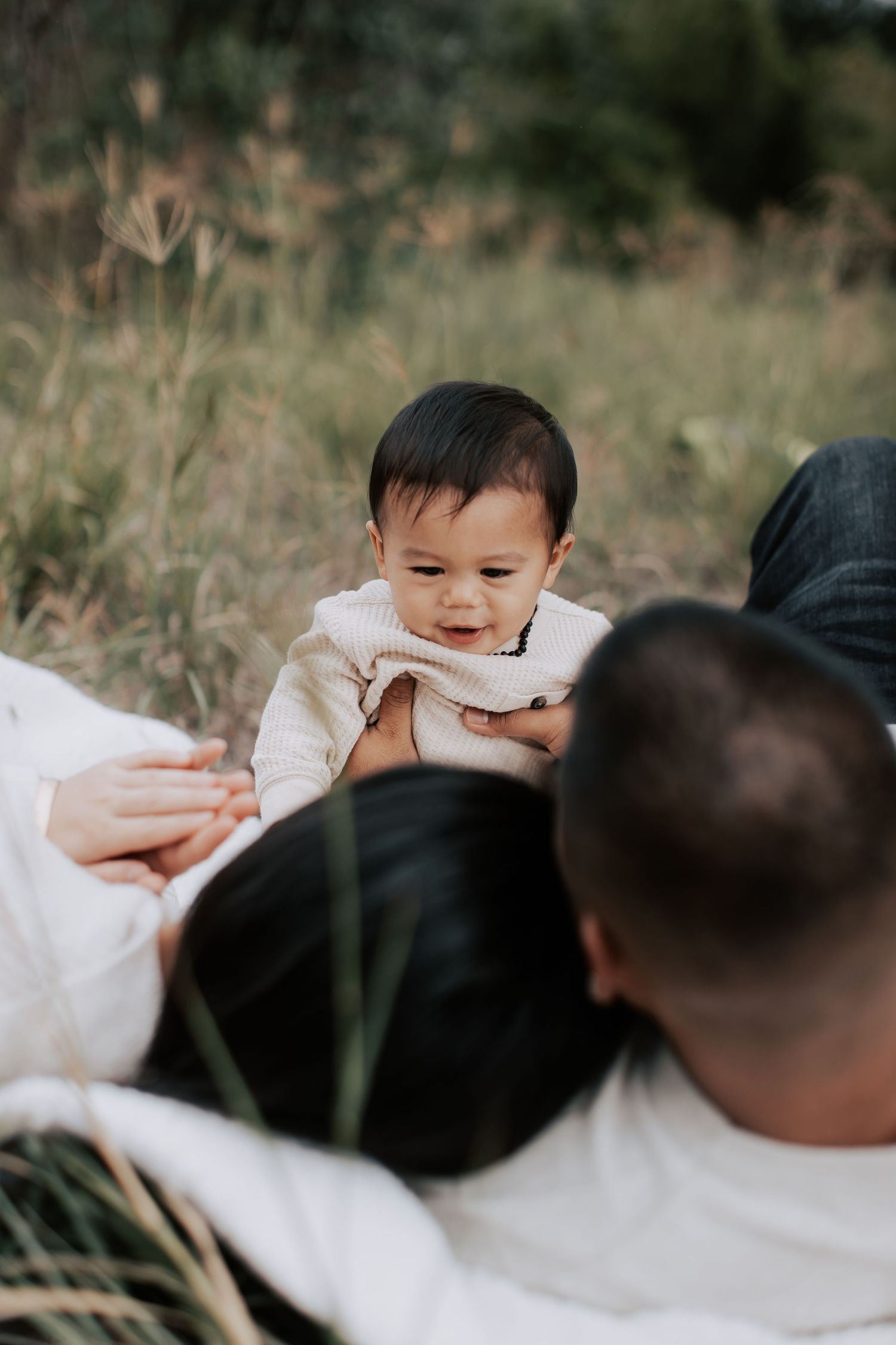 Baby sitting between parents lying in a grassy field, wearing a beige outfit, smiling softly outdoors.
