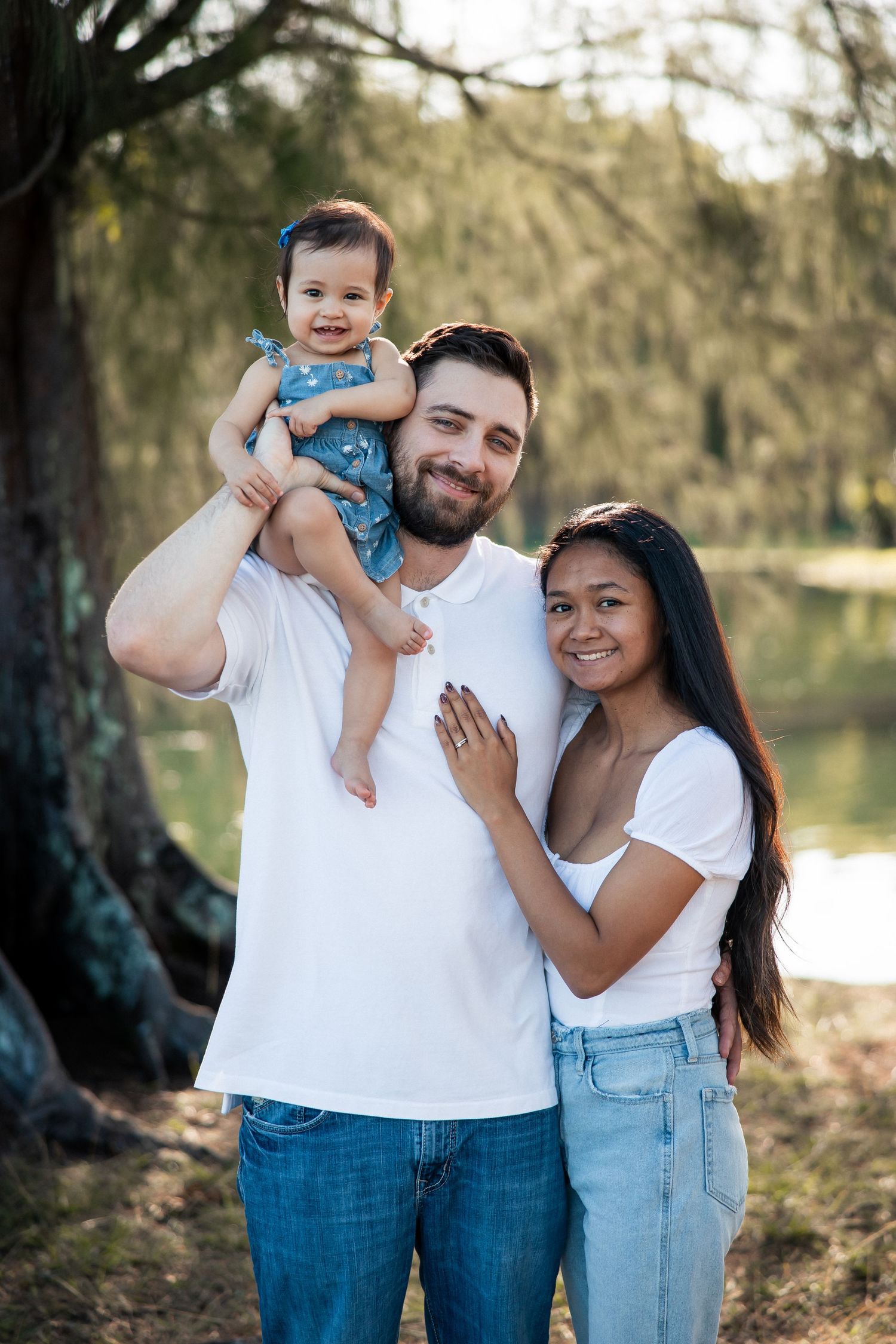 Happy interracial family of three posing outdoors near a lake, father holding baby girl, mother embracing him.