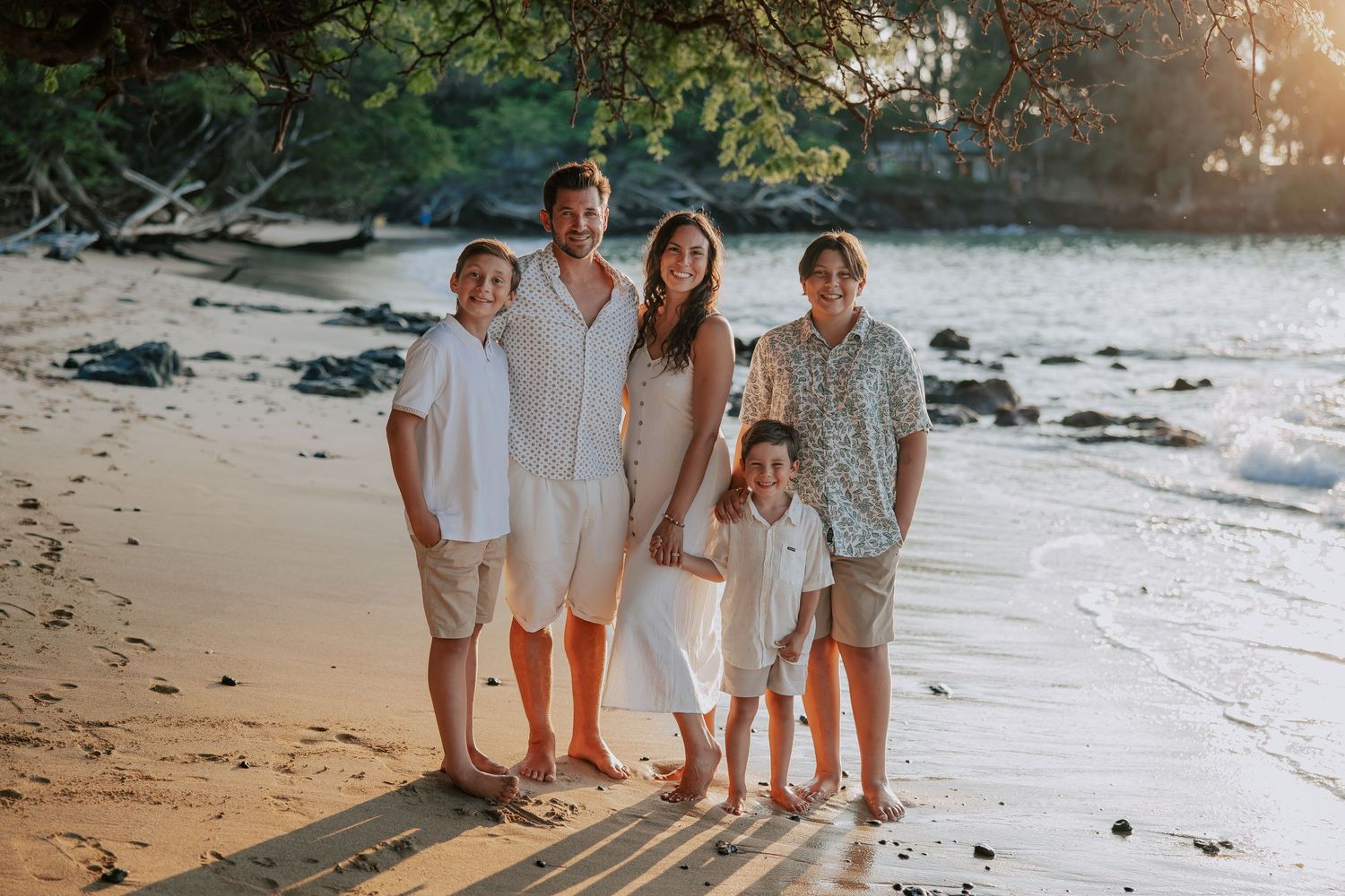 Family of five smiling together on a tropical beach at golden hour, wearing white and neutral outfits.
