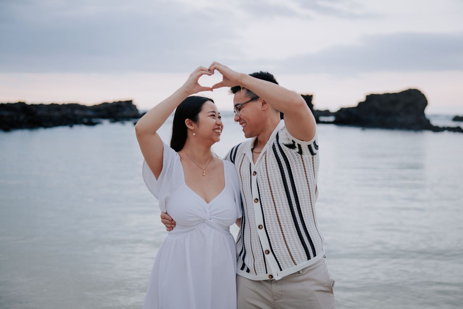 Couple making heart shape with hands at beach, romantic seaside photo shoot with ocean backdrop.