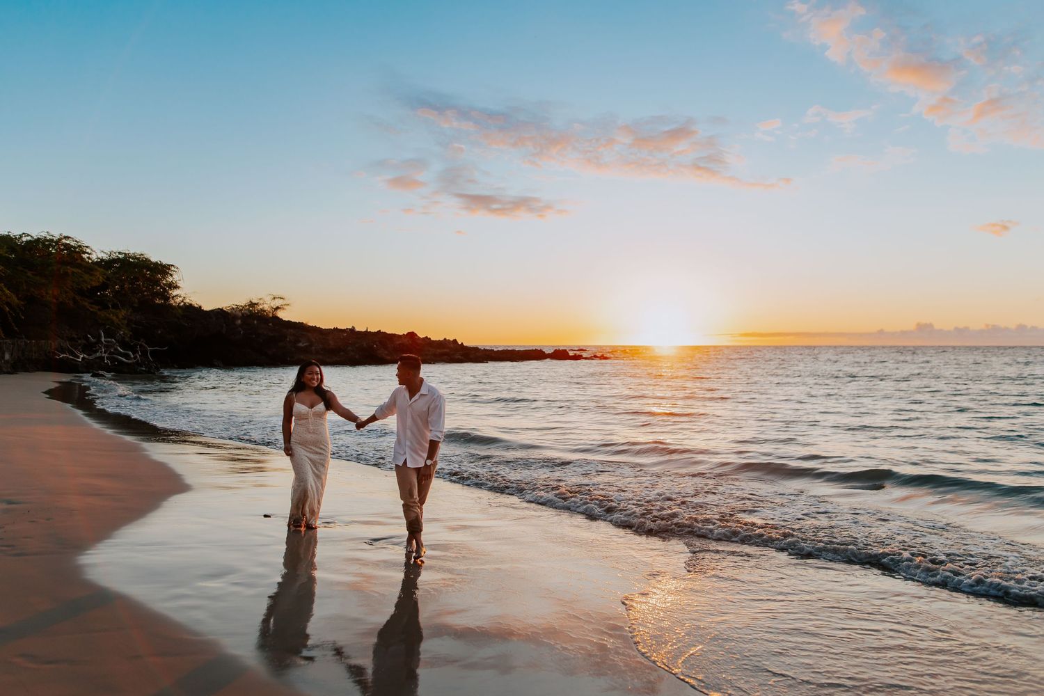 Couple holding hands walking on a tropical beach at sunset with golden sky and gentle waves.