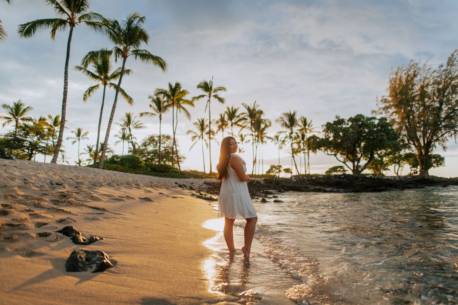 Woman in white dress walking on sandy Hawaiian beach at sunset with tropical palm trees in background.