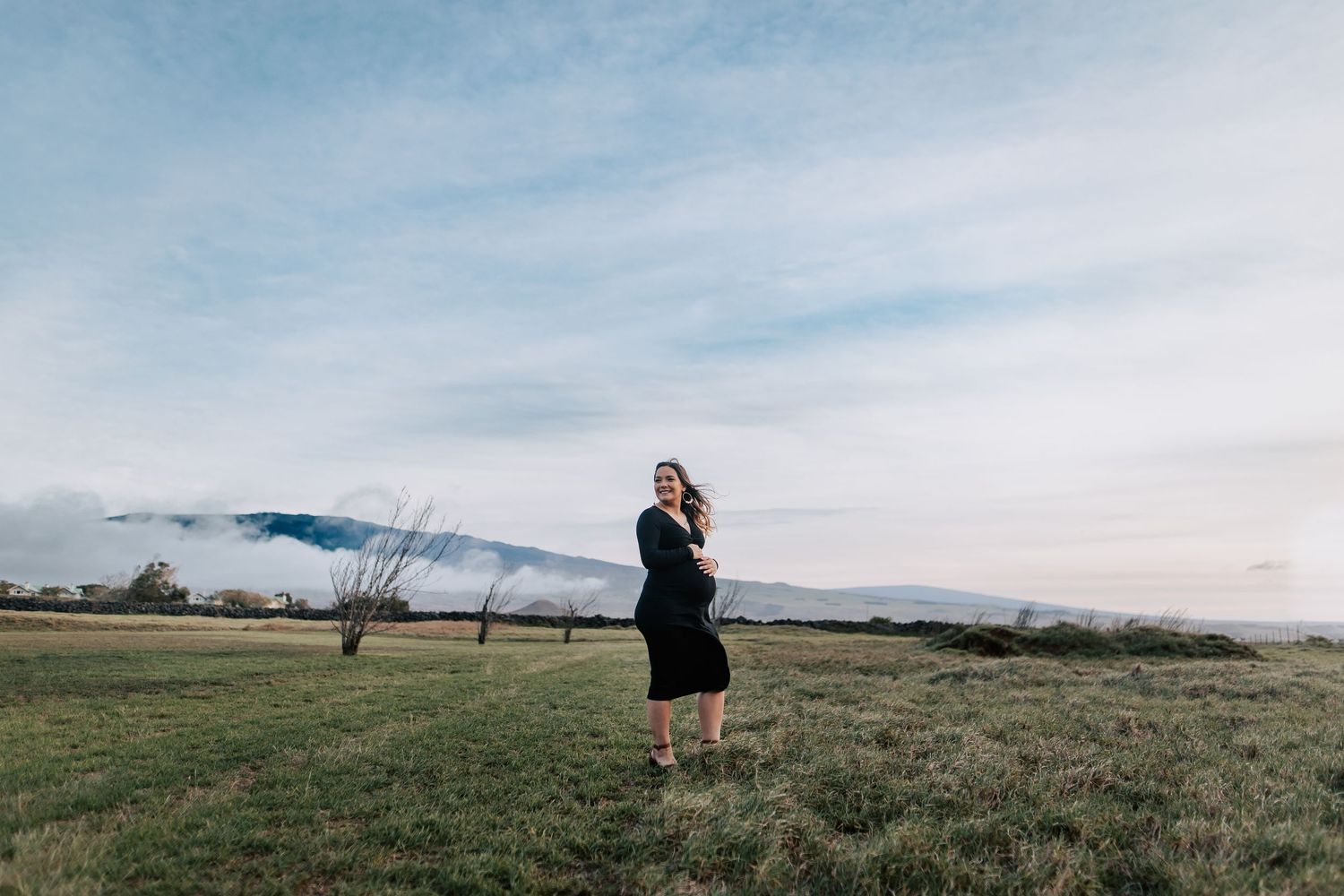 Pregnant woman in black dress standing in open field with mountain backdrop and cloudy sky.