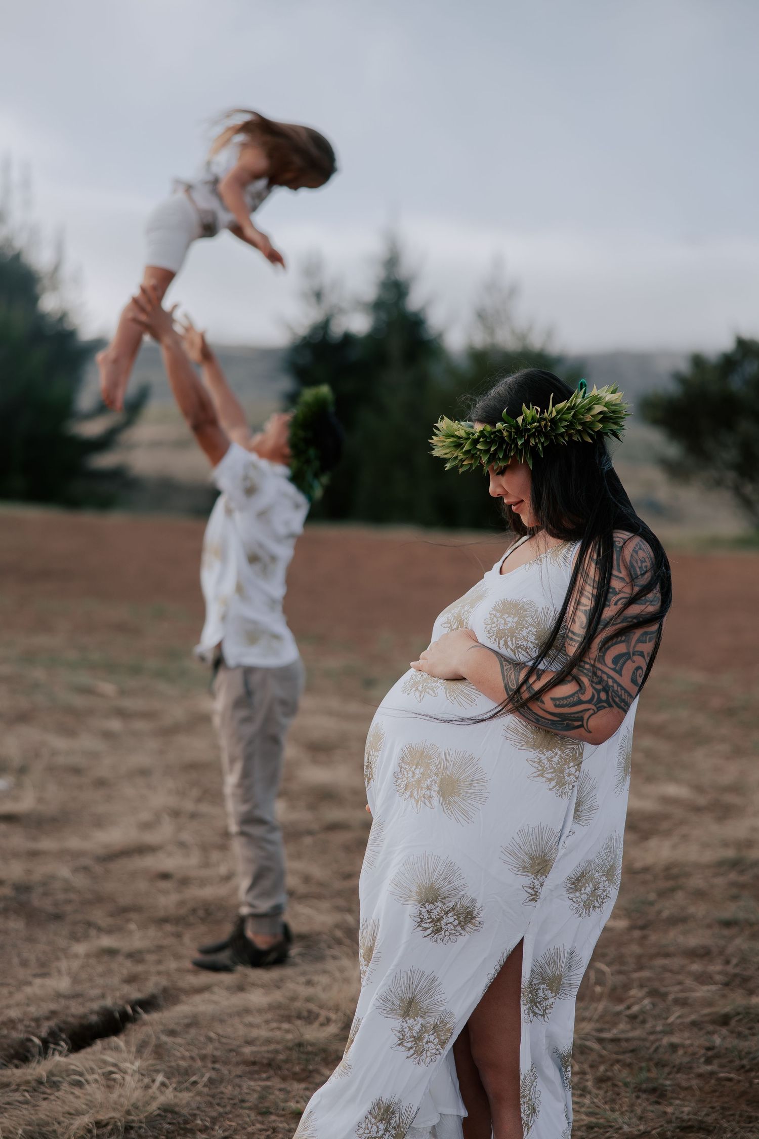 Pregnant woman in floral crown and white dress stands in field while man tosses child in air behind her.