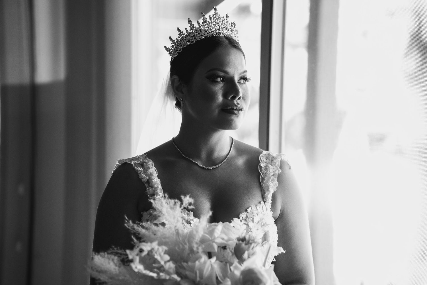 Black and white portrait of a bride wearing a tiara and floral wedding gown, gazing thoughtfully out a window.