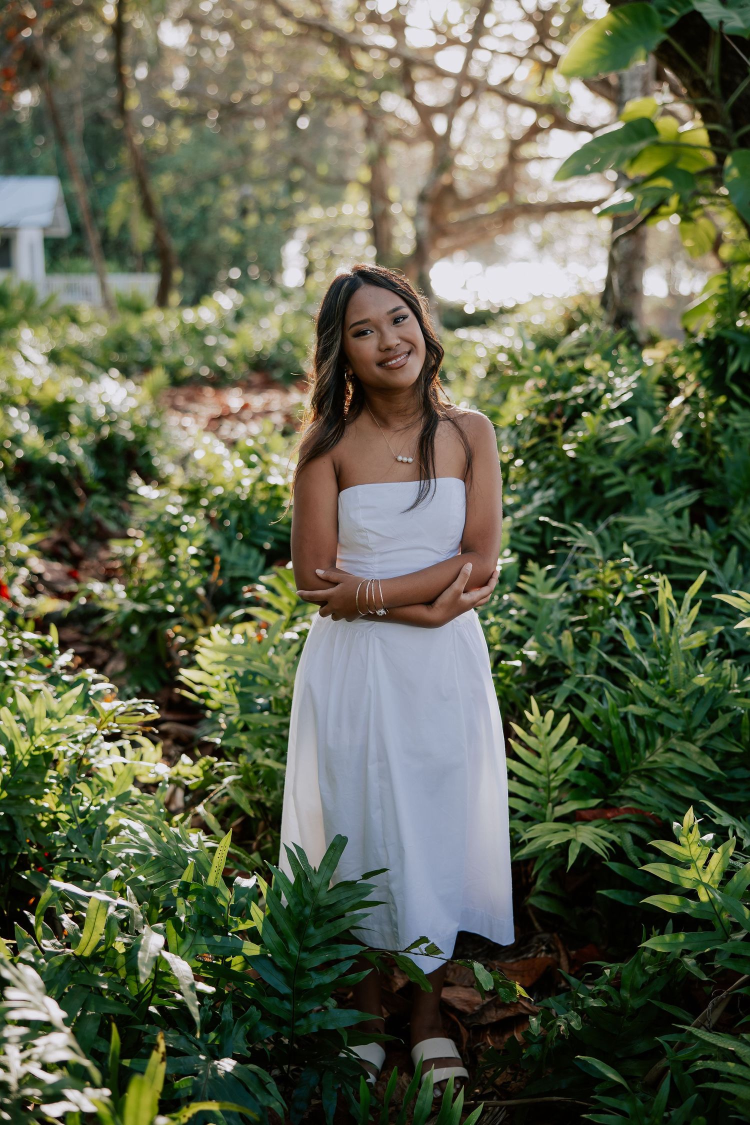 Young woman in white strapless dress smiling in lush green garden surrounded by tropical ferns and foliage.