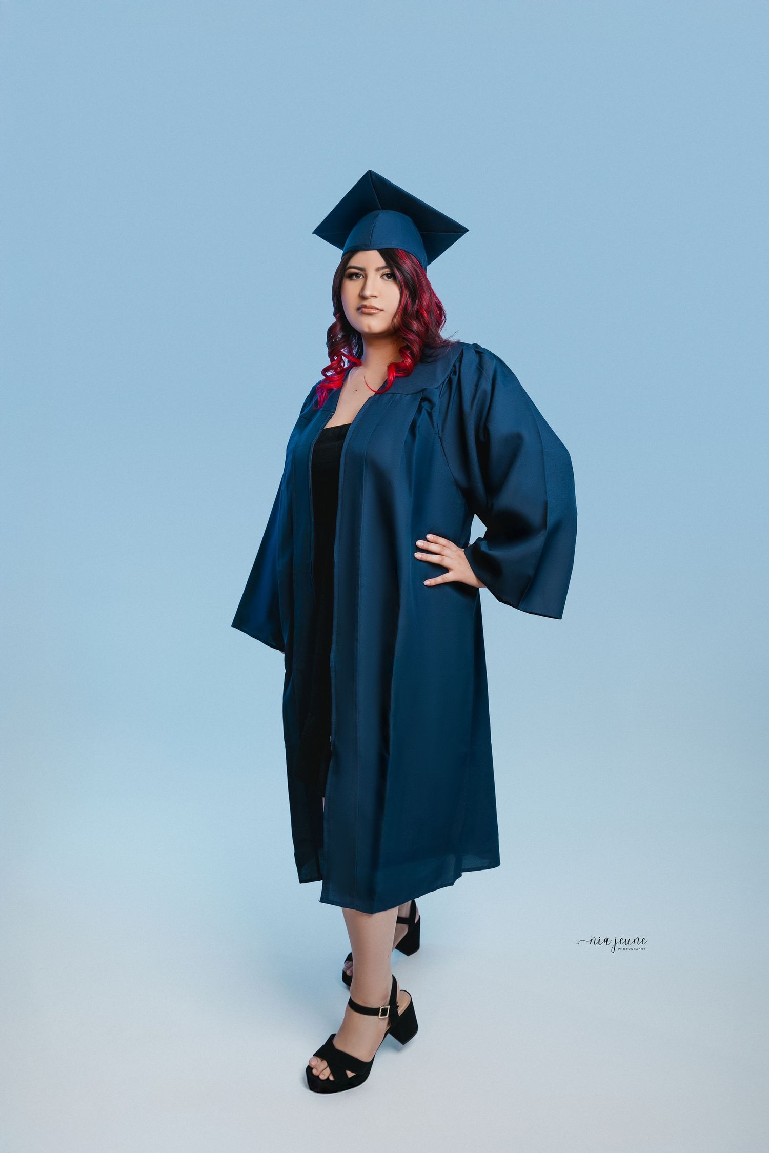 Graduate in navy blue cap and gown poses confidently against light blue background.